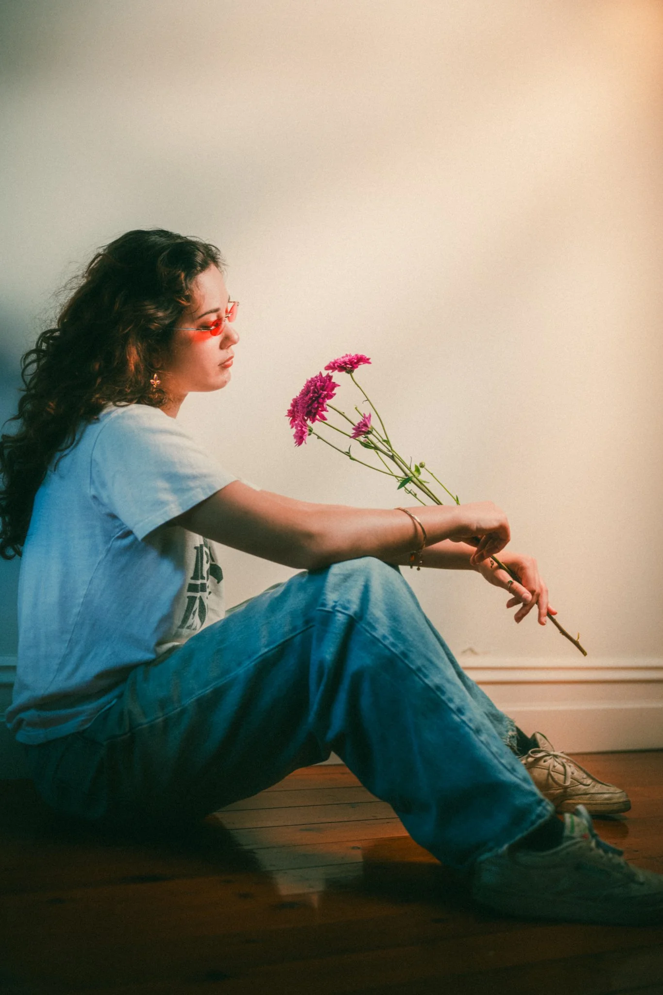 A woman sitting on a wooden floor against a plain wall, holding a pink flower bouquet. She has wavy hair, wears red-tinted sunglasses, a white t-shirt, loose blue jeans, sneakers, and has accessories like bracelets.