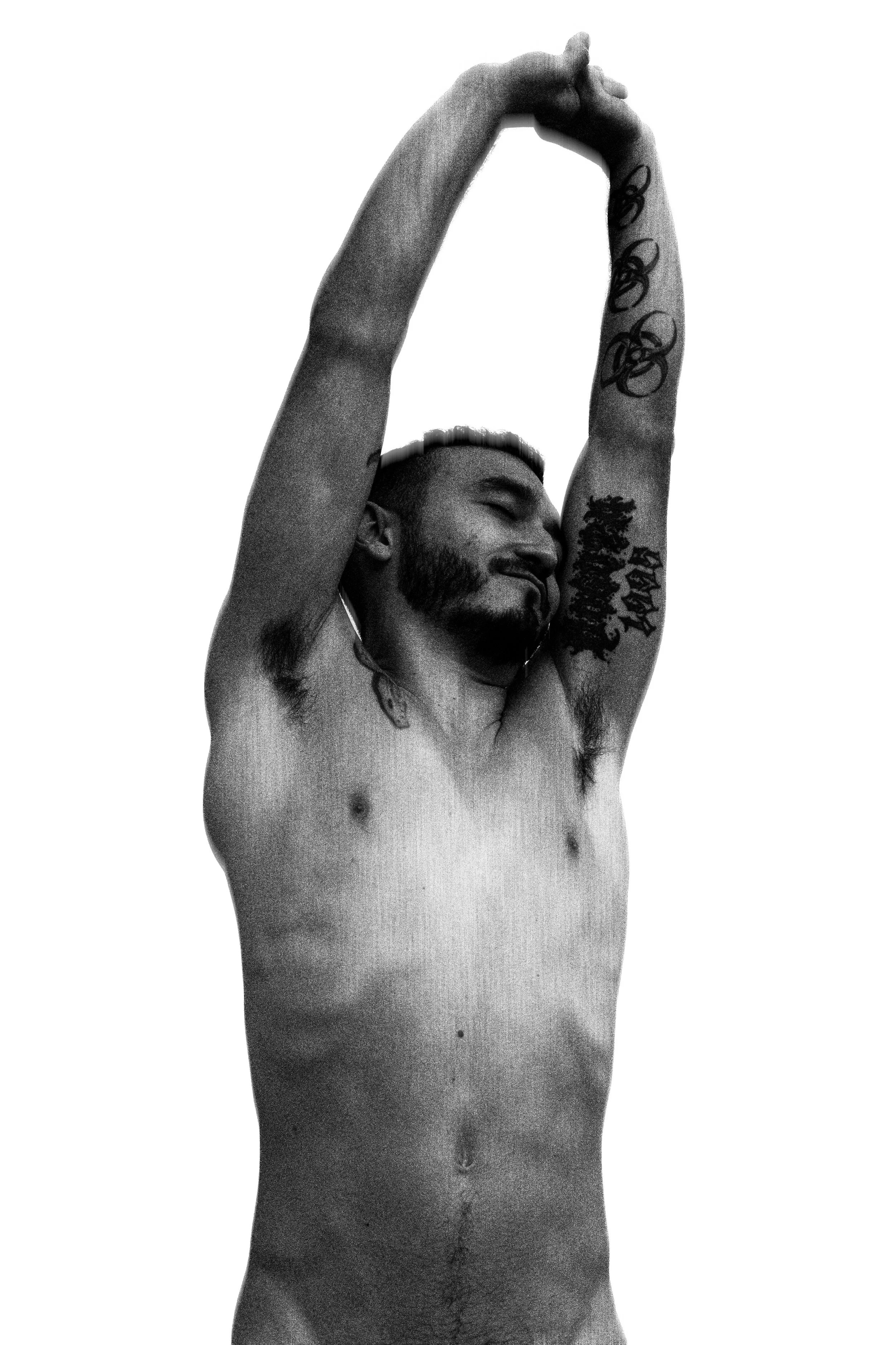 A shirtless man with tattoos stretching his arms above his head against a plain background.