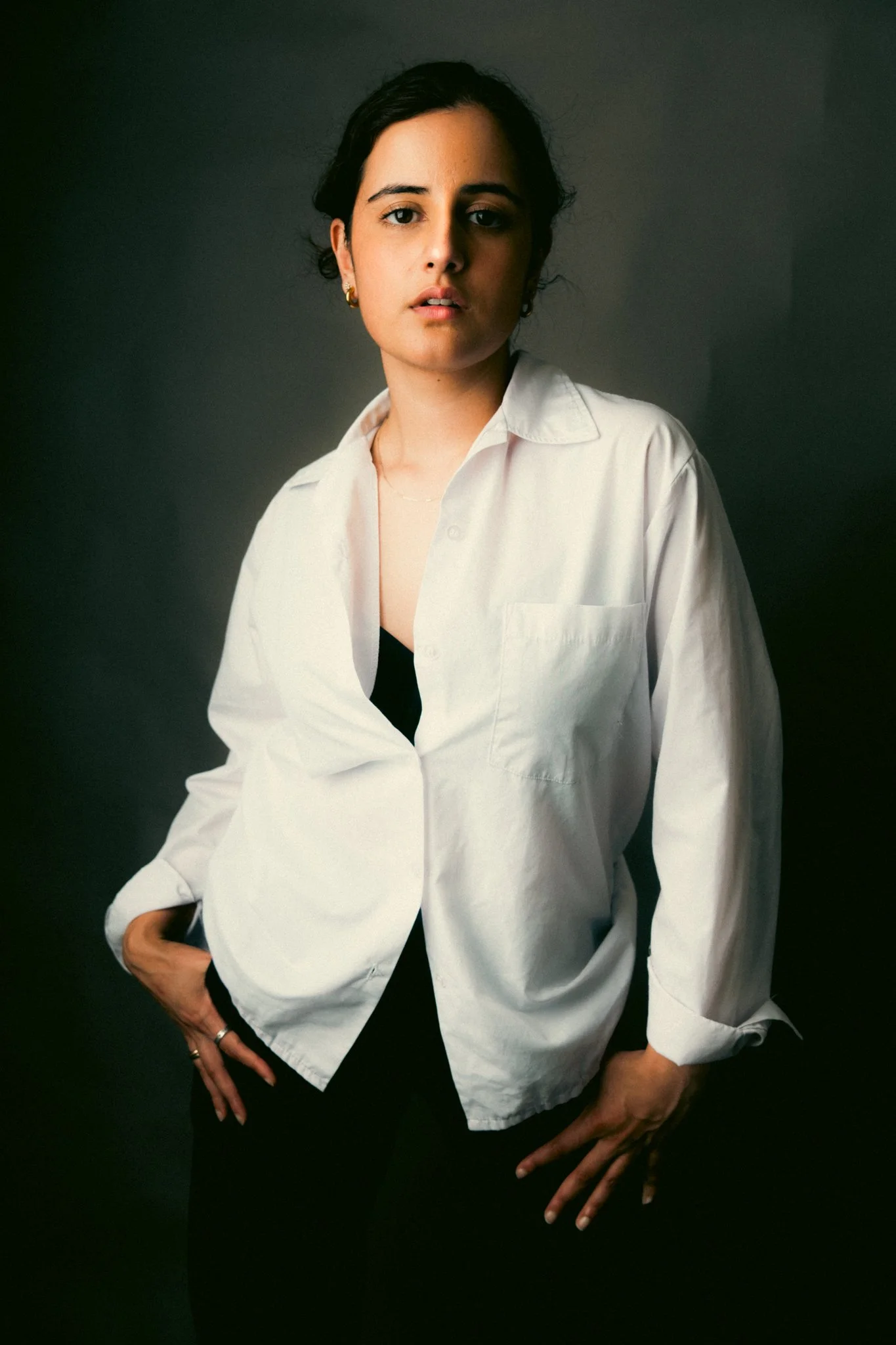 A woman with dark hair wearing a white shirt, black outfit, and gold earrings, standing against a dark background.