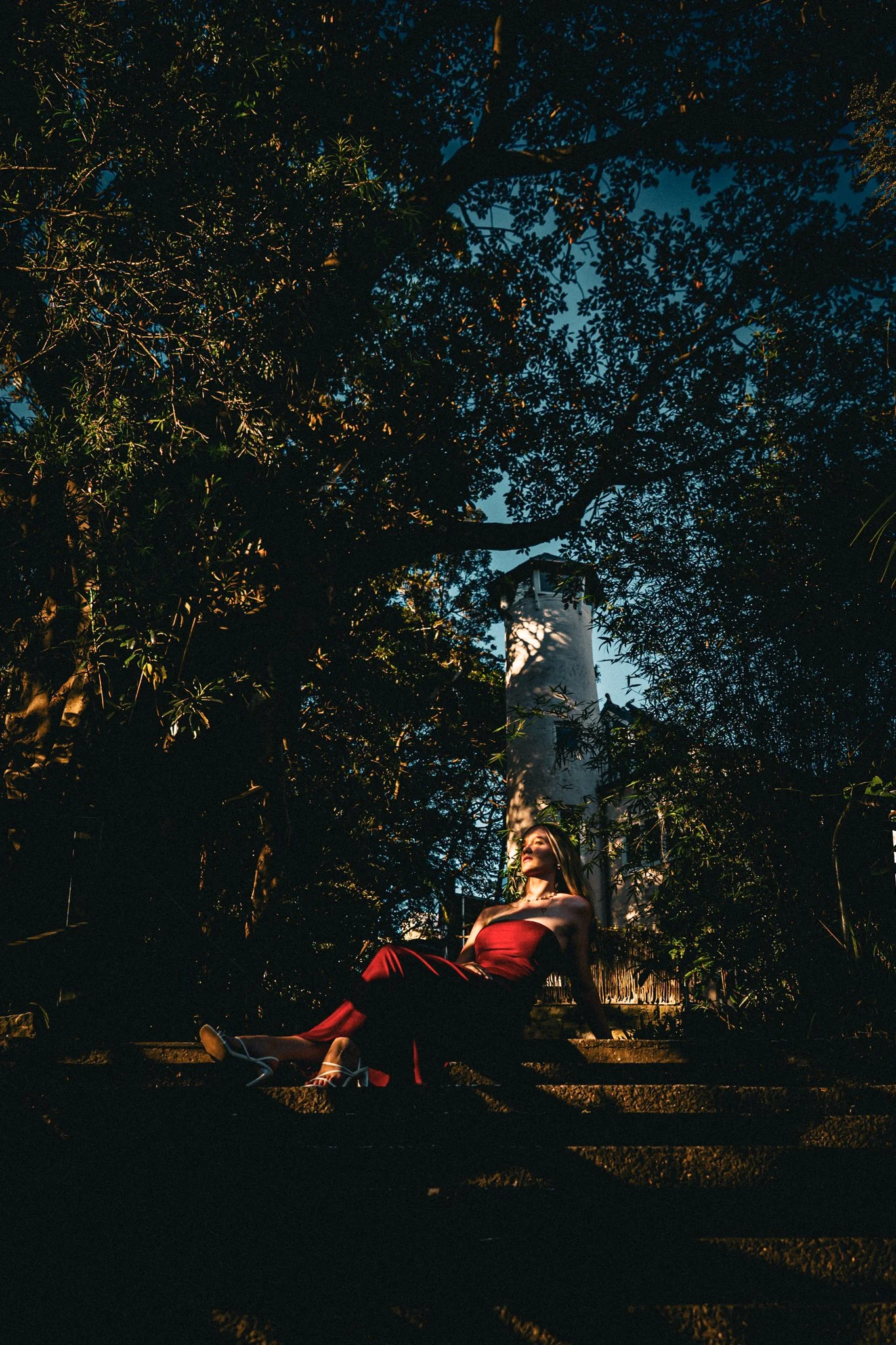 A woman in a red strapless dress and white heels sitting on outdoor steps at sunset, surrounded by trees with a lighthouse in the background.