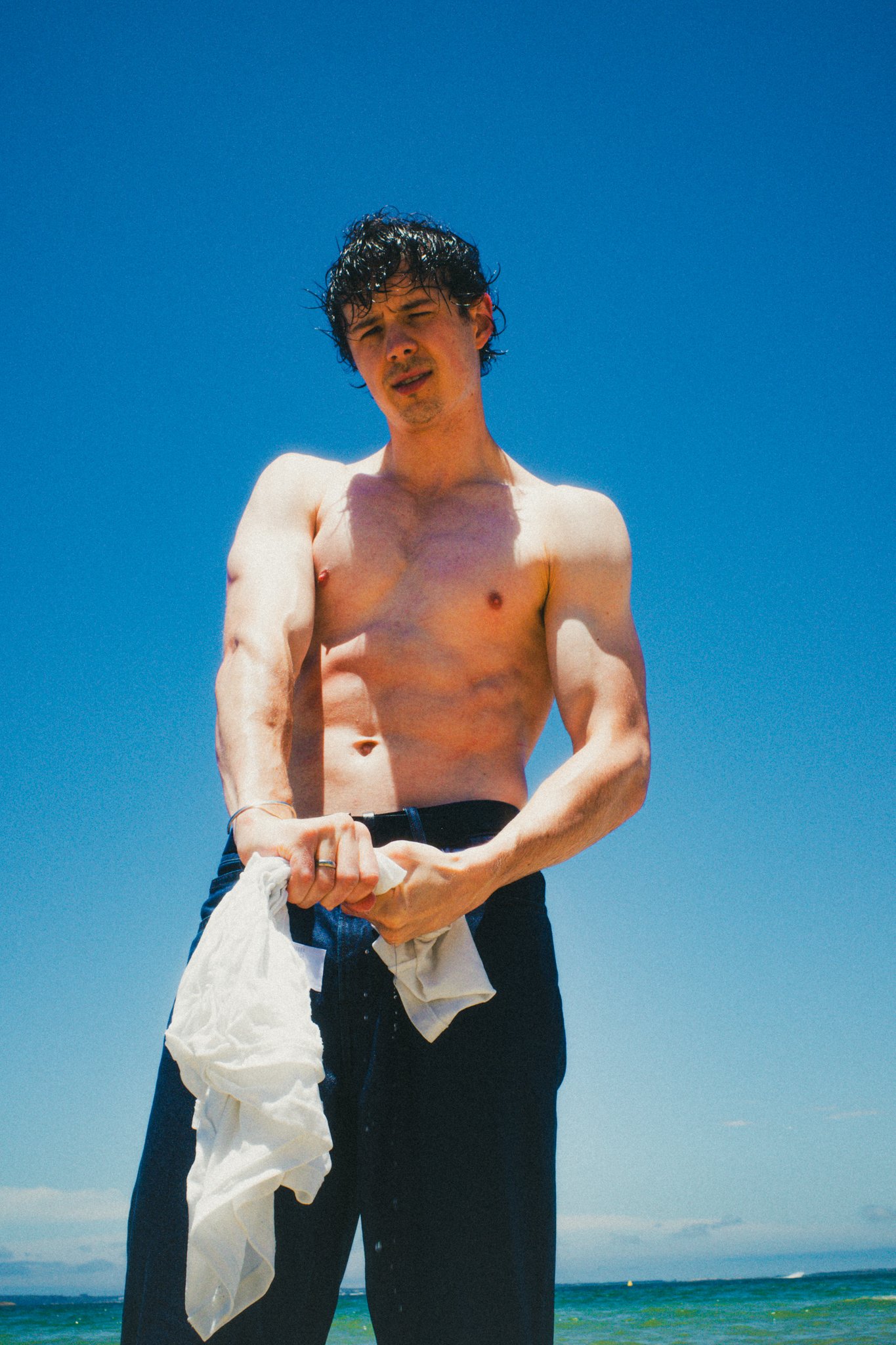 A shirtless man holding a white towel, standing outdoors under a clear blue sky near the ocean.