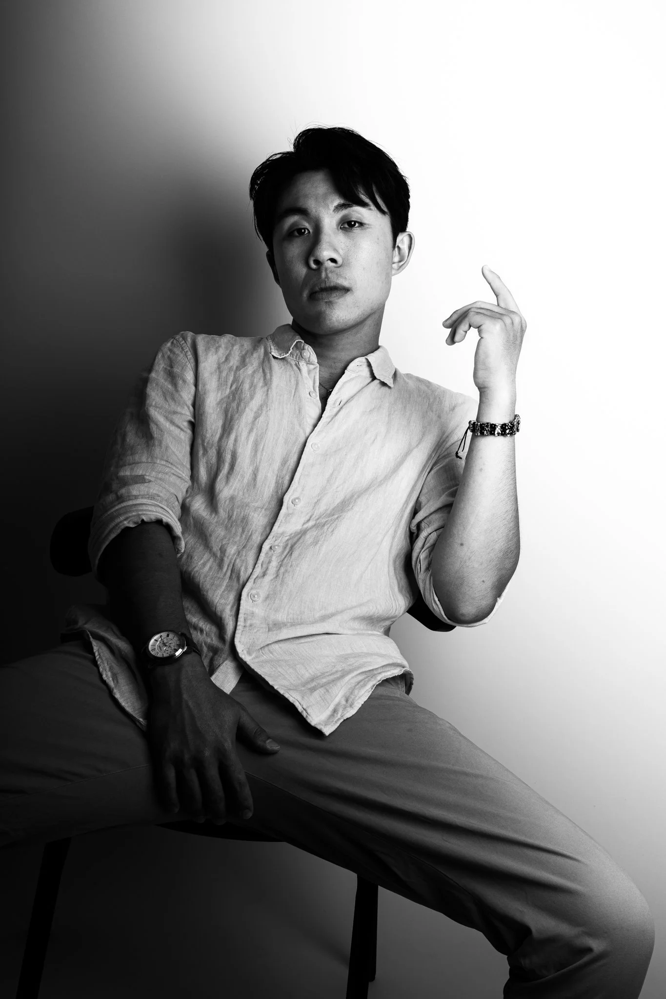 Black and white portrait of a young man sitting on a chair, wearing a button-up shirt and pants, with one arm resting on his knee and the other hand raised near his head, looking directly at the camera.