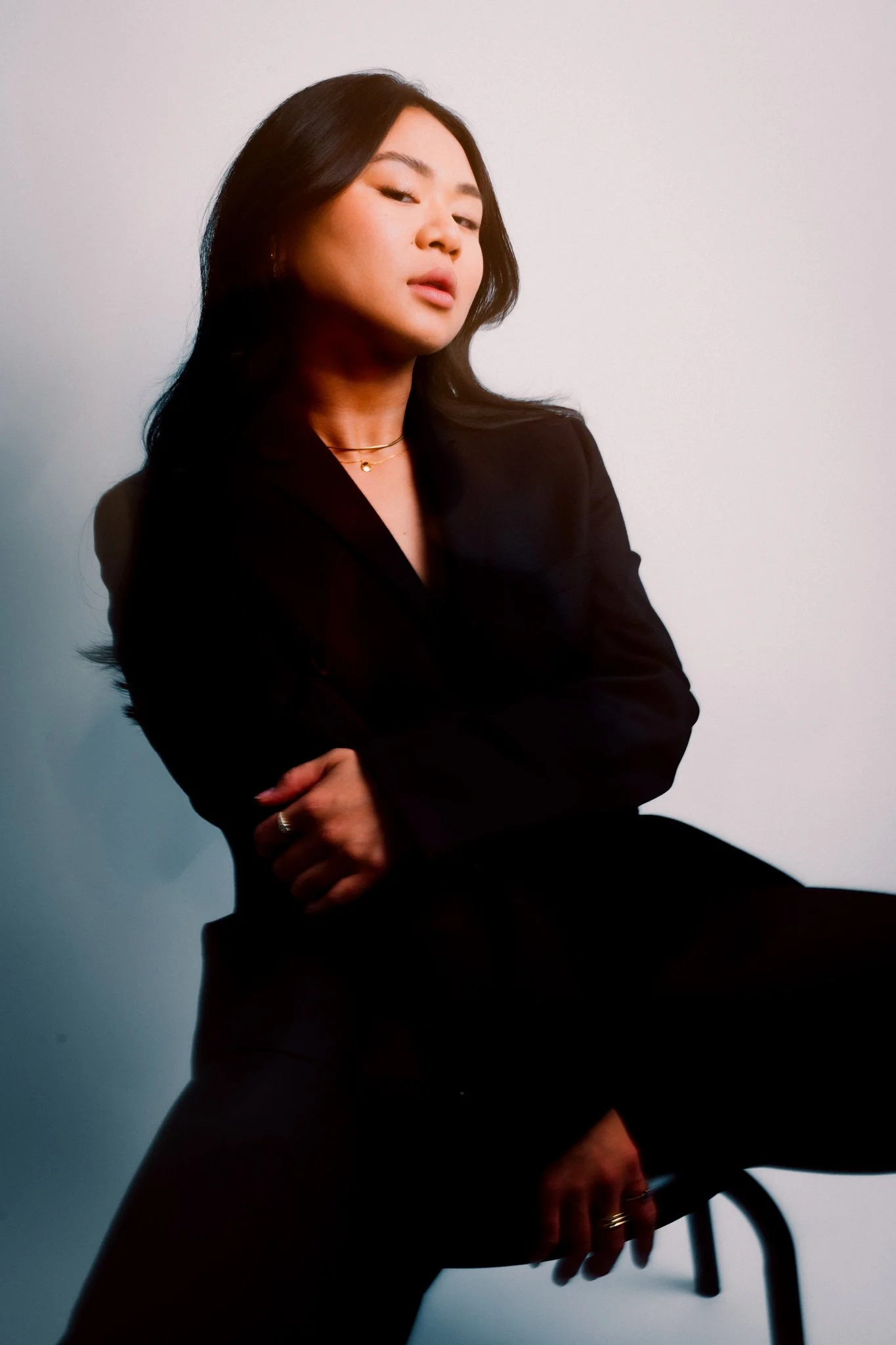 A woman with long black hair wearing a black blazer and jewelry, posing against a plain wall.