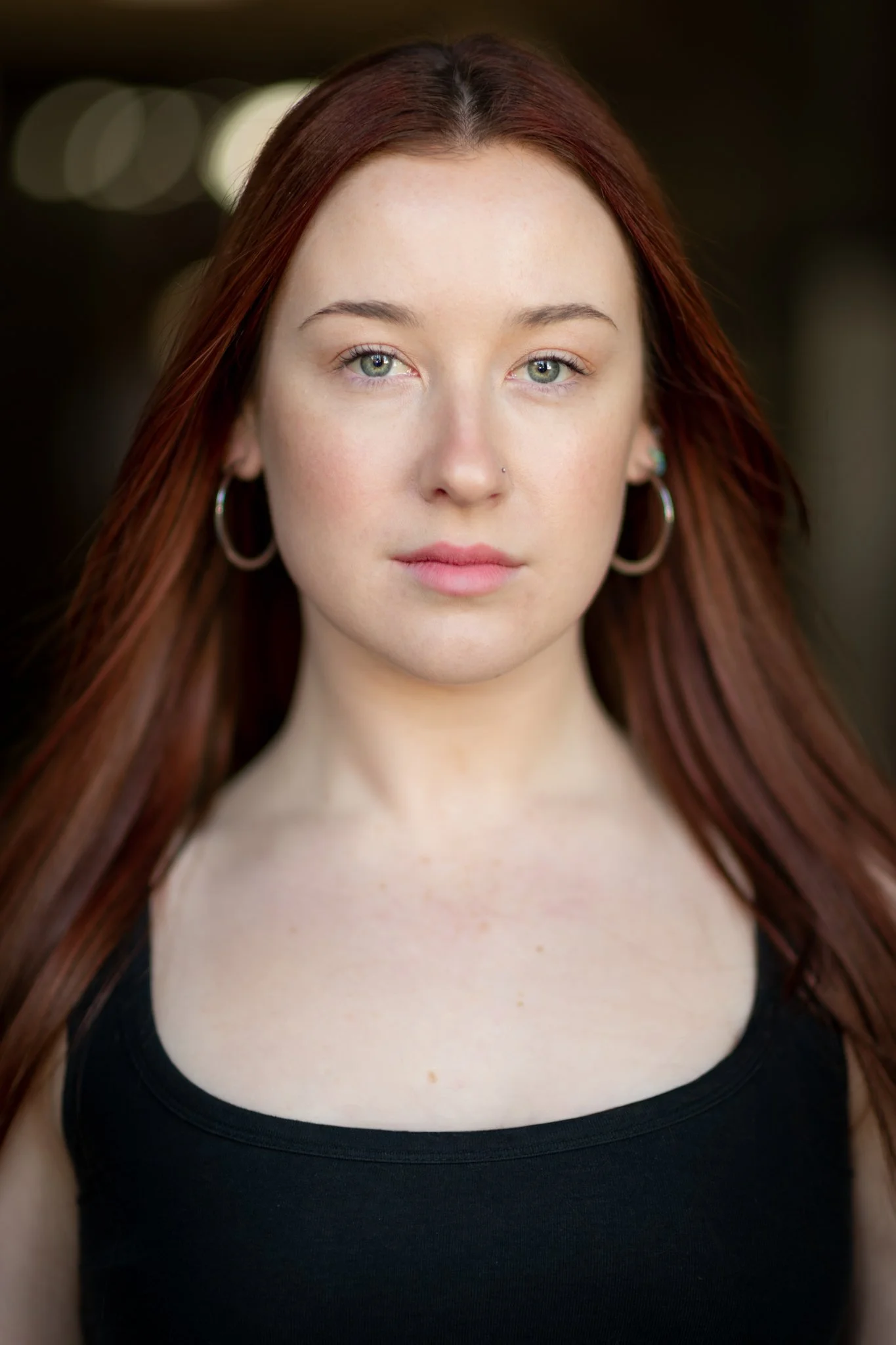 Close-up portrait of a young woman with red hair, wearing hoop earrings and a black sleeveless top, looking directly into the camera with a neutral expression.