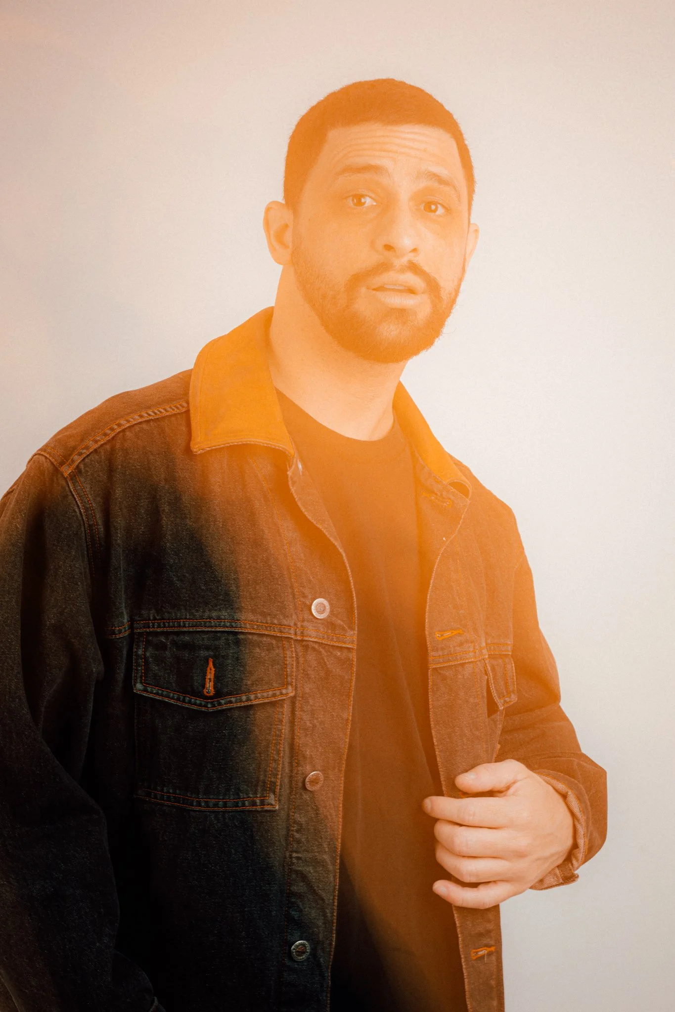 A man with short dark hair and a beard wearing a black shirt and a brown denim jacket, standing against a plain white background.