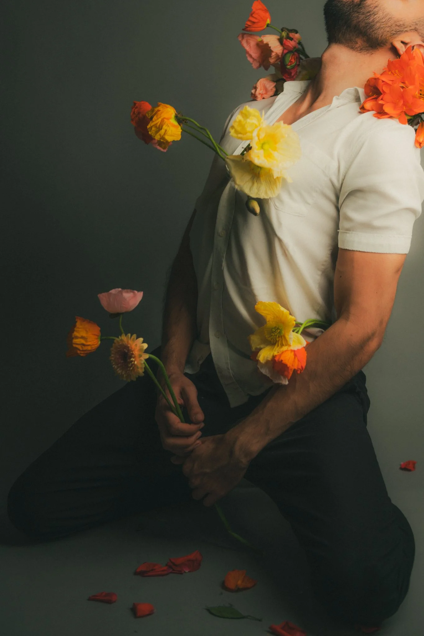 A man wearing a white shirt and black pants is kneeling on the floor, surrounded by colorful flowers and flower petals. The flowers are placed on his body and held in his hand, with some petals scattered on the ground.
