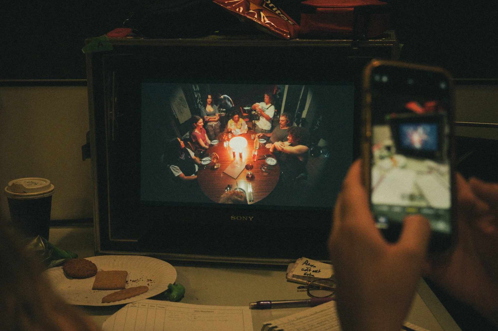 A person is taking a photo of a group of people sitting around a table with a glowing lamp on it, displayed on a Sony monitor. The image includes a plate with food, a cup, and some papers and snacks on the table.