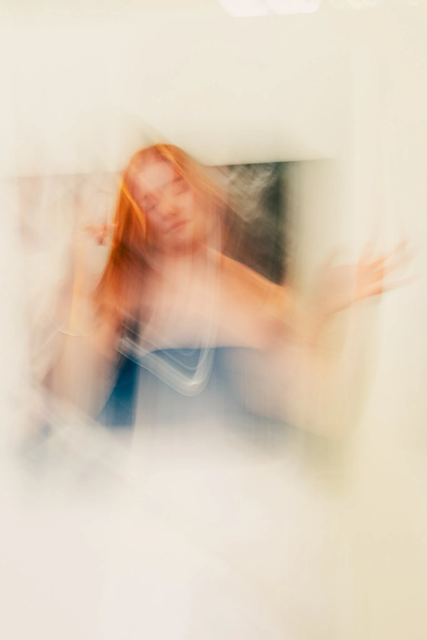 Blurred image of a woman with red hair posing with one hand on her face and the other extended, wearing a blue top, against a plain background.