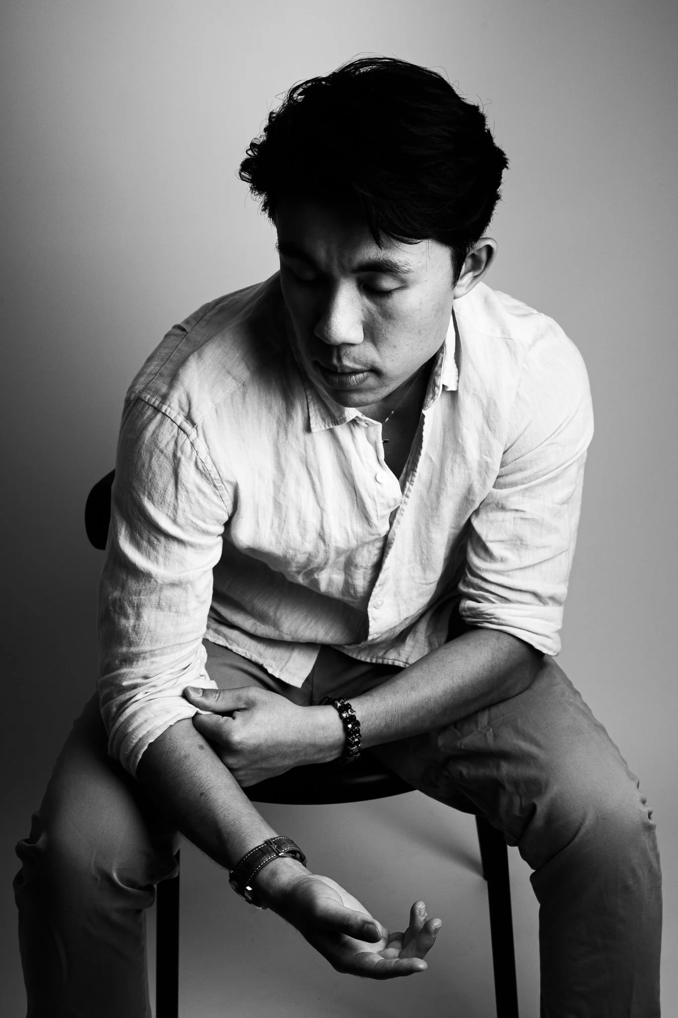 A black and white photograph of a young man sitting on a chair with his eyes closed, wearing a light-colored shirt rolled up at the sleeves, pants, wristwatch, and bracelets.