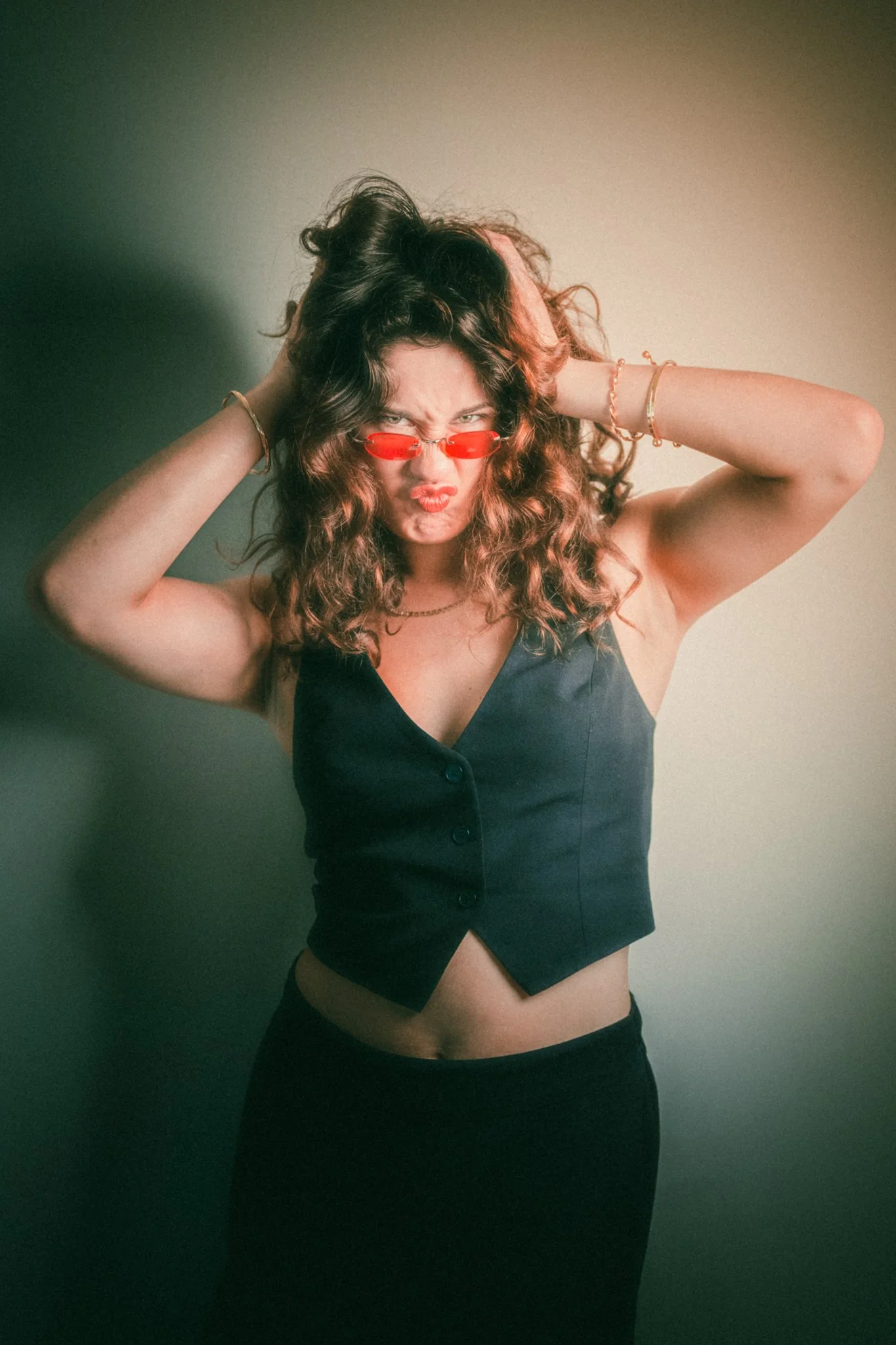 A woman with curly hair wearing red-tinted sunglasses, a black vest, and black skirt, making a fierce facial expression with pursed lips.