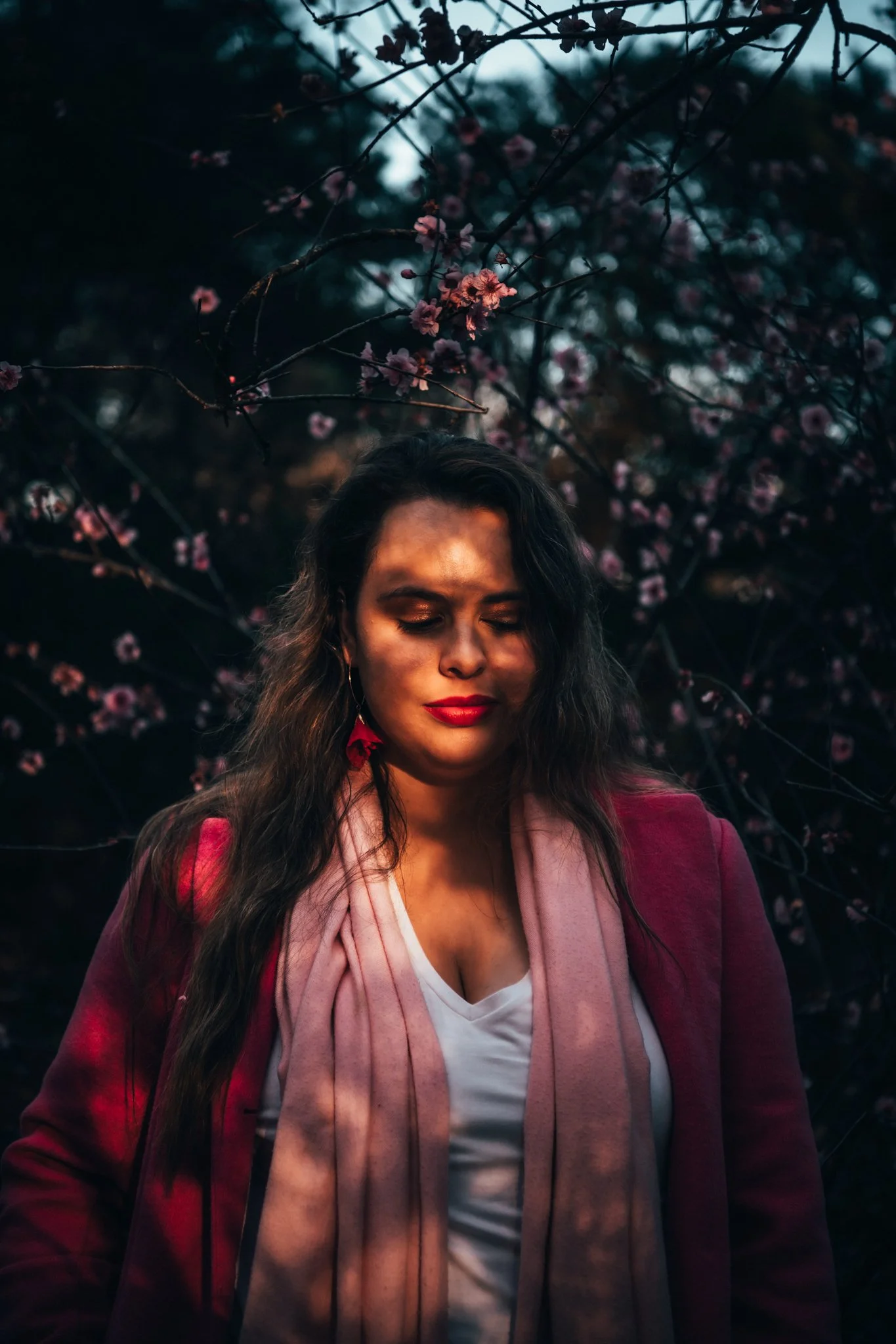 A woman with long dark hair and red lipstick stands outdoors with pink blossoms in the background. She is wearing a red blazer, a white top, and a pink scarf, with her eyes closed.
