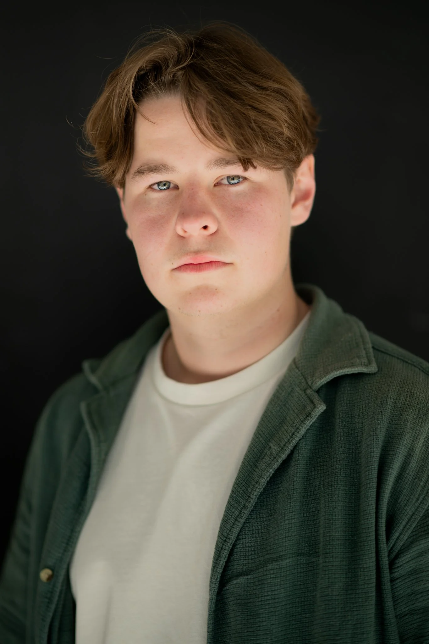 Headshot of a young man with light skin, brown hair, and blue eyes, wearing a white t-shirt and a green jacket, standing in front of a black background.