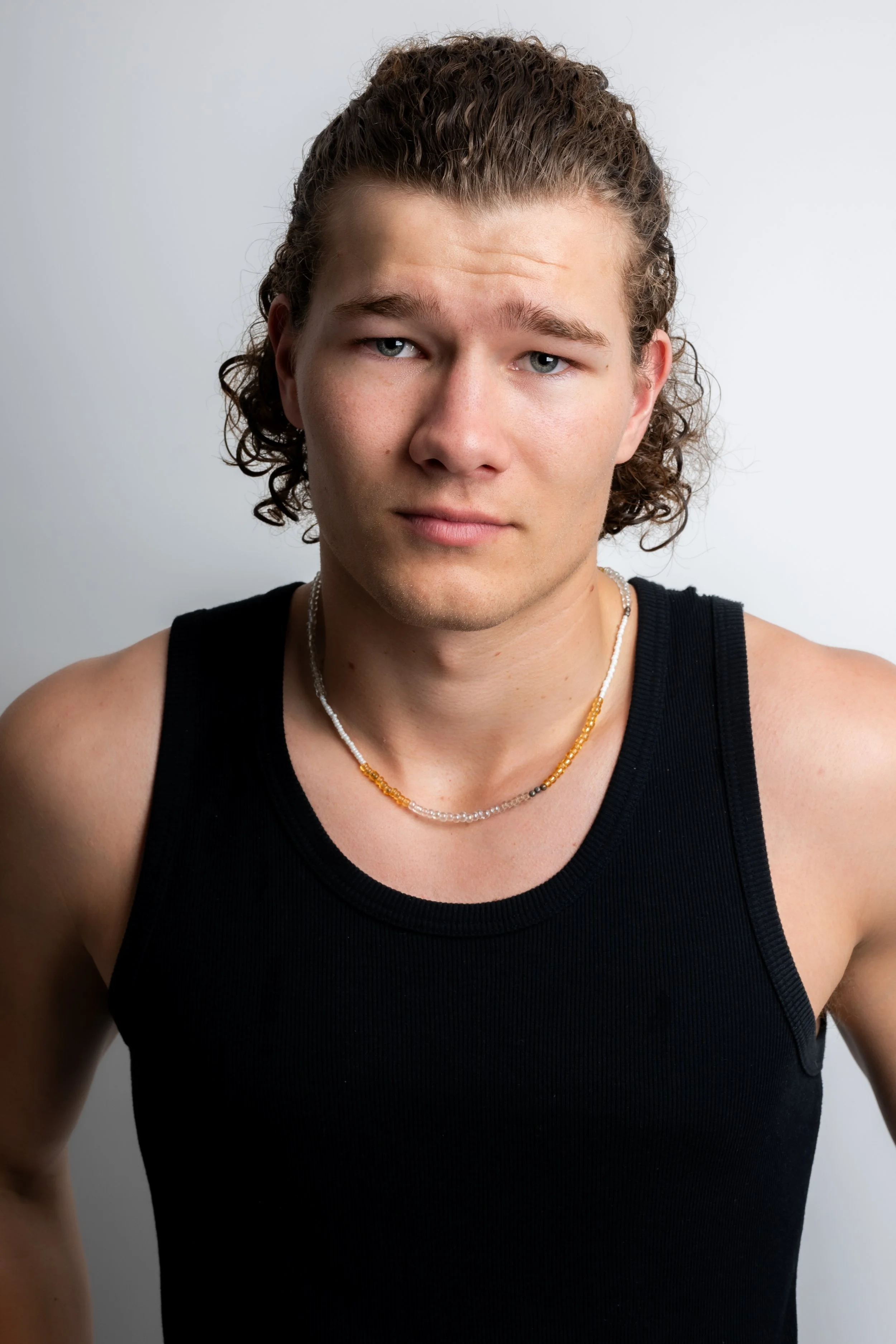 A young man with curly hair wearing a black sleeveless shirt and beaded necklaces, looking directly at the camera with a neutral expression.
