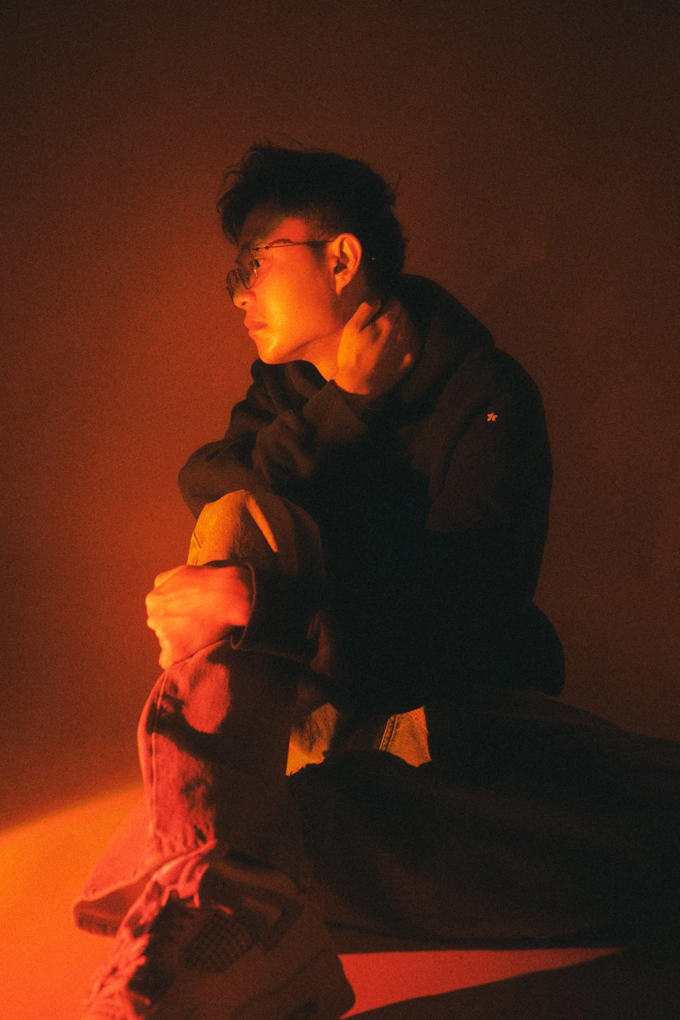A young man with glasses, wearing a black hoodie and ripped jeans, sitting on the floor in low, reddish lighting, gazing to the side with one hand on his neck.