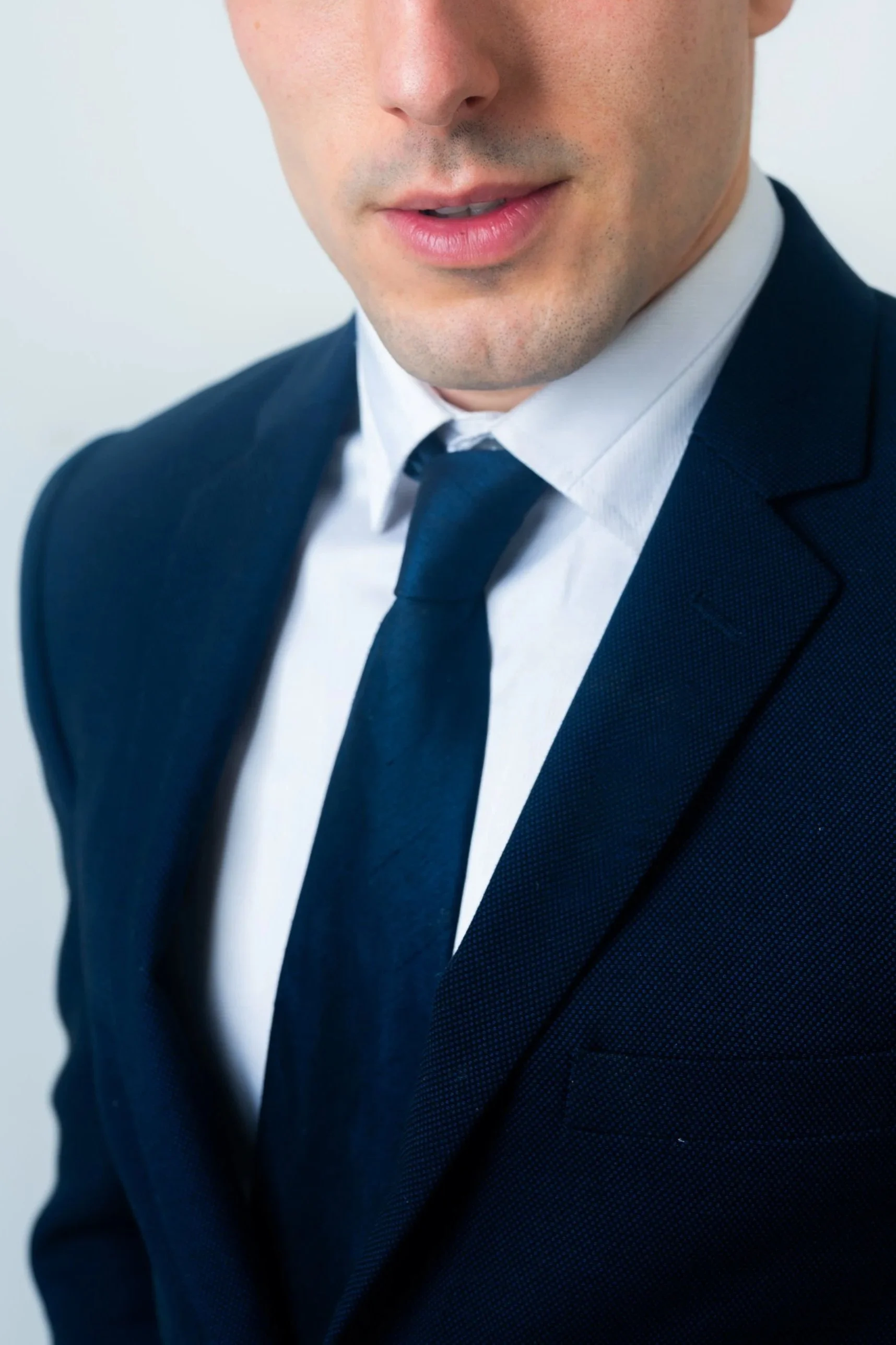 Close-up of a man wearing a navy blue suit, white shirt, and navy tie, with the lower half of his face visible.