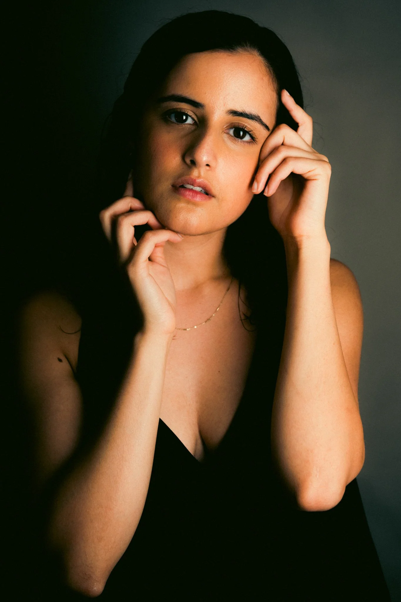 Portrait of a woman with dark hair, touching her face and neck, wearing a black top and a delicate gold necklace, against a dark background.