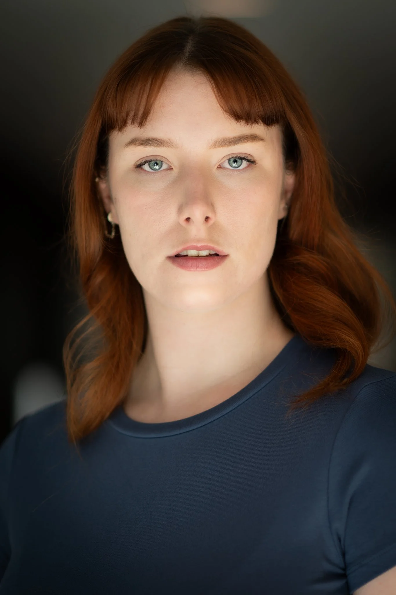 Close-up portrait of a young woman with red hair and blue eyes wearing a navy blue shirt, looking at the camera against a dark background.