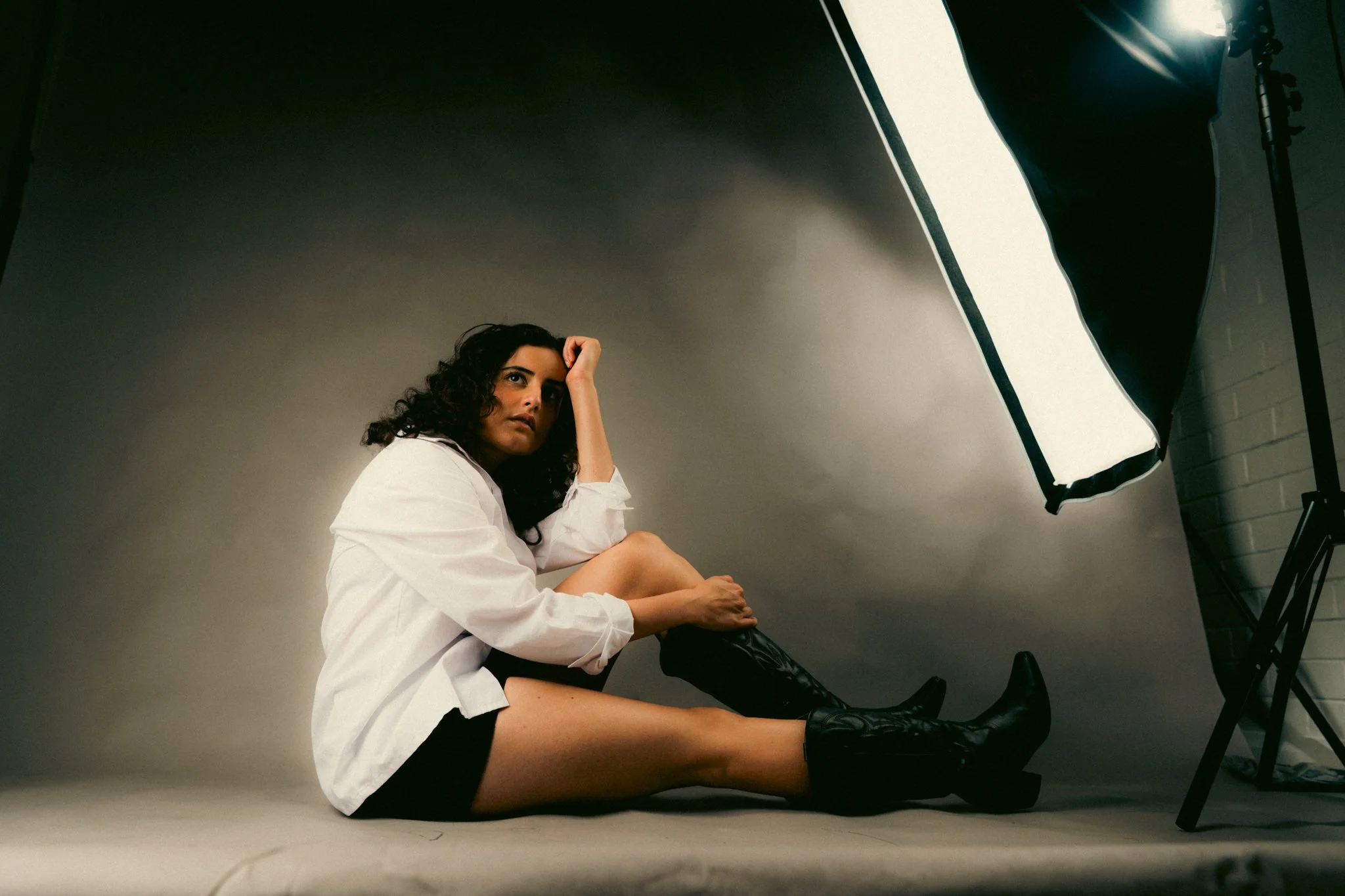 A woman sitting on the floor in a photography studio, wearing a white shirt and black boots, with a large studio light on a stand directed at her.