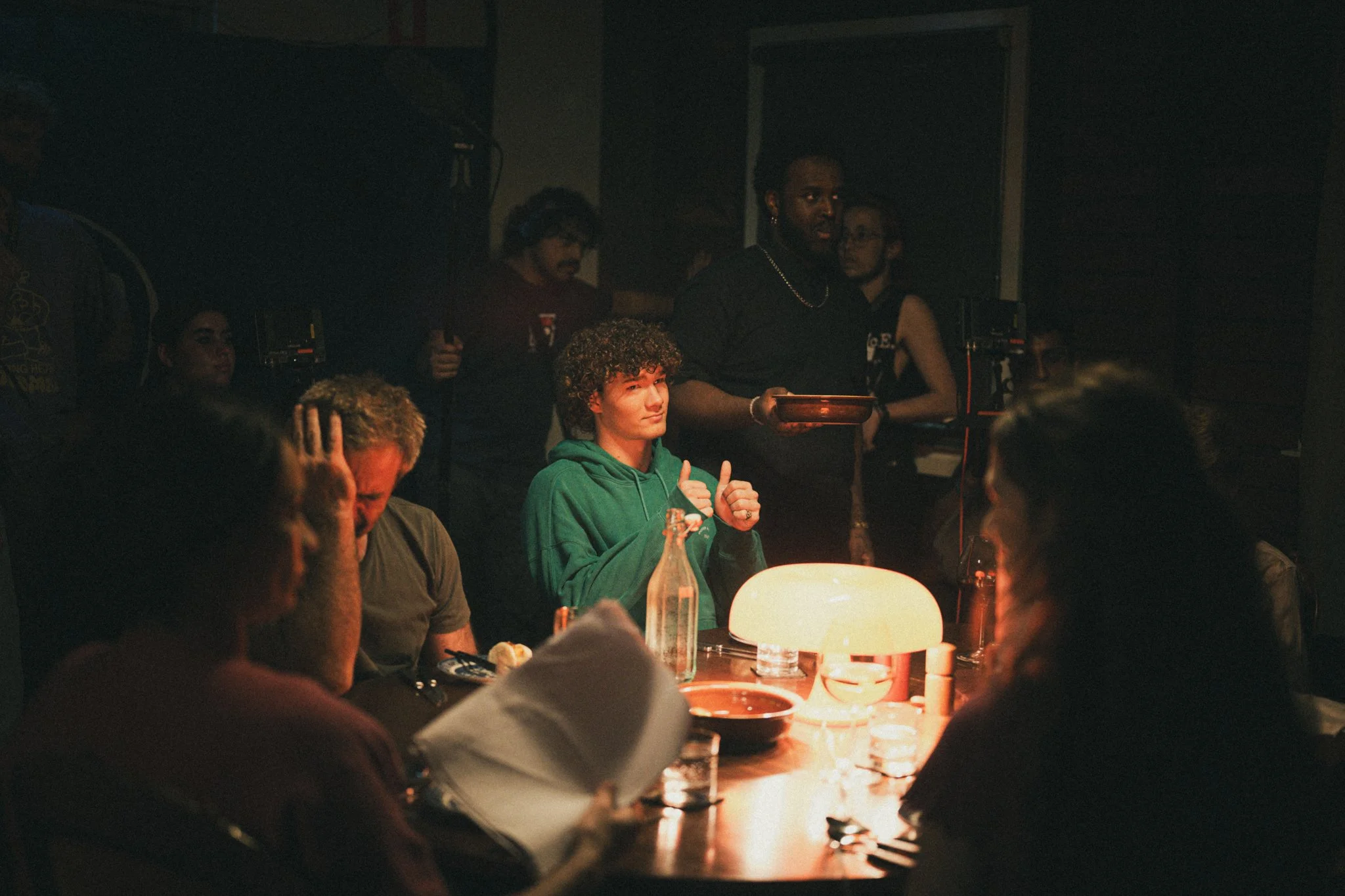 A young man in a green hoodie giving a thumbs up at a dimly lit dinner table with several people. An illuminated lamp is on the table. In the background, team members and a staff member holding a tray are present.