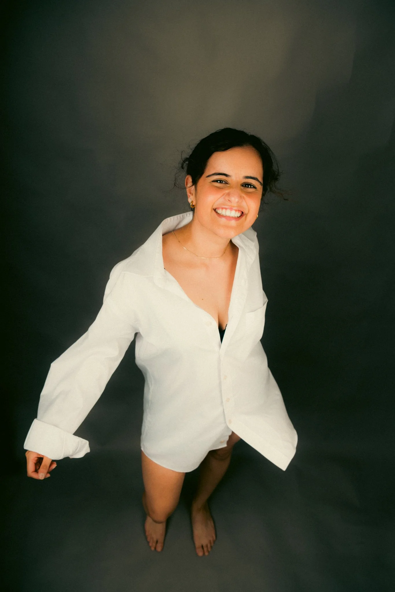 Young woman with dark hair, smiling, wearing an oversized white shirt, standing barefoot on a dark background.