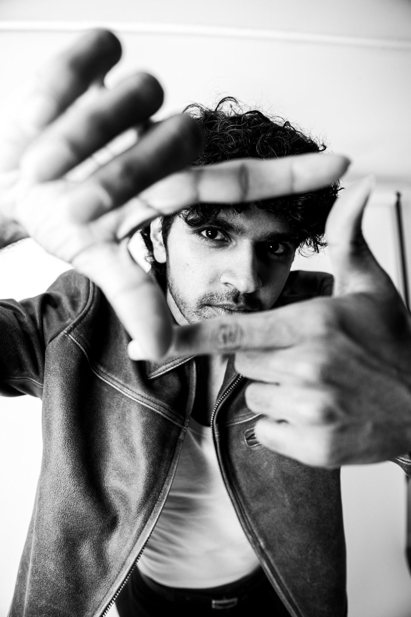Black and white photo of a man with curly hair in a leather jacket, looking directly at the camera through a frame made with his fingers.