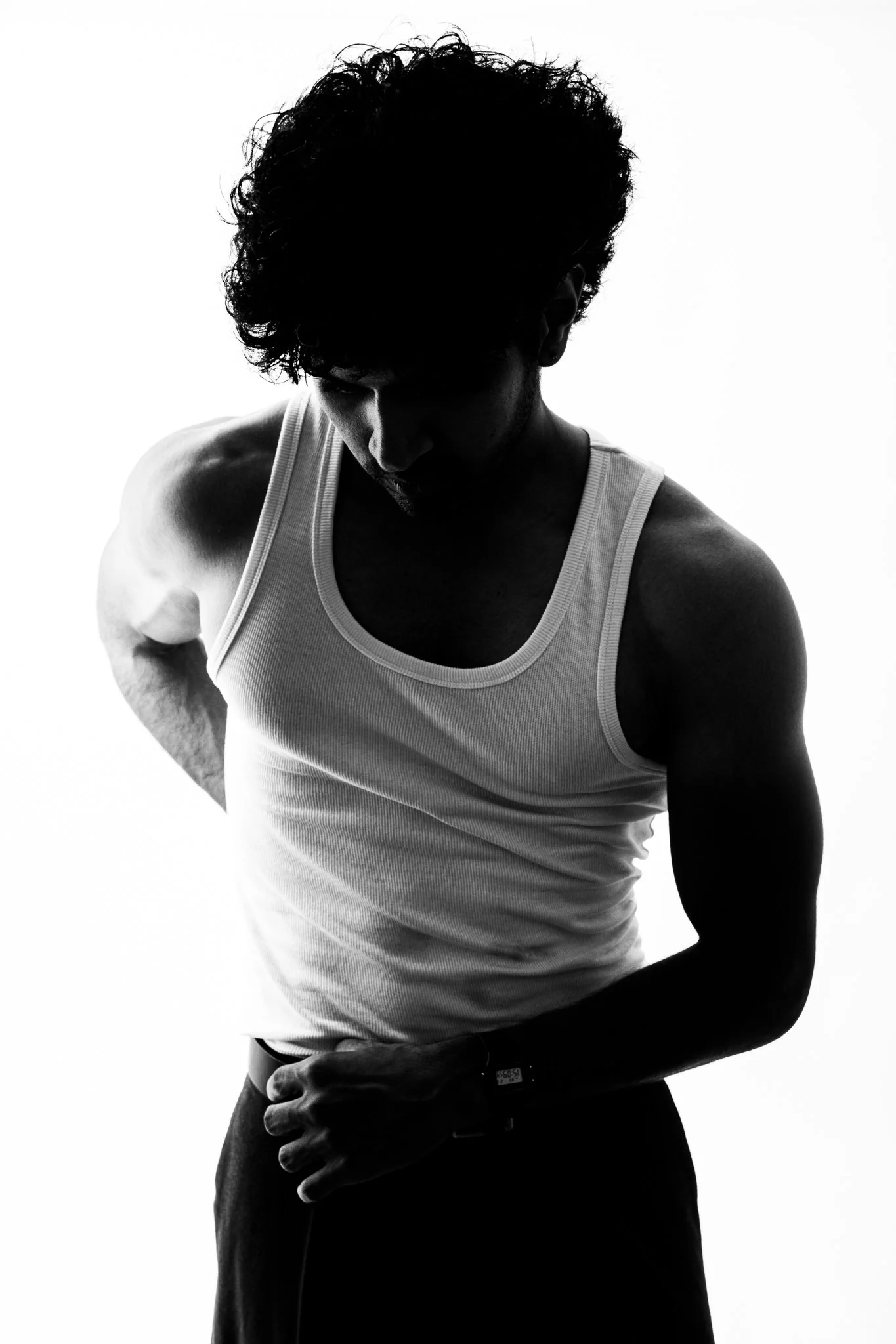 A silhouette of a man wearing a sleeveless shirt, looking downward with his hand near his waist against a bright background.