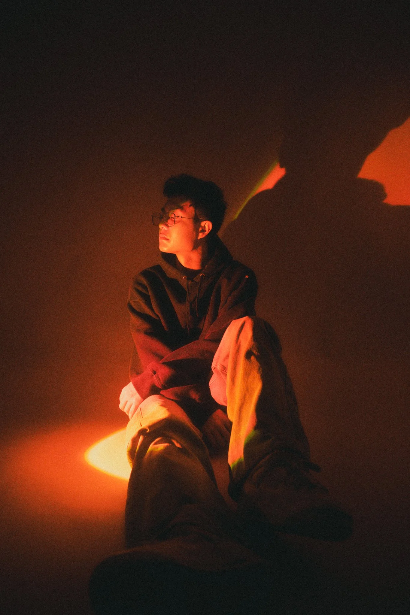 A young man sitting on the floor in a dark room illuminated by orange and yellow lighting, with shadows cast on the wall behind him.