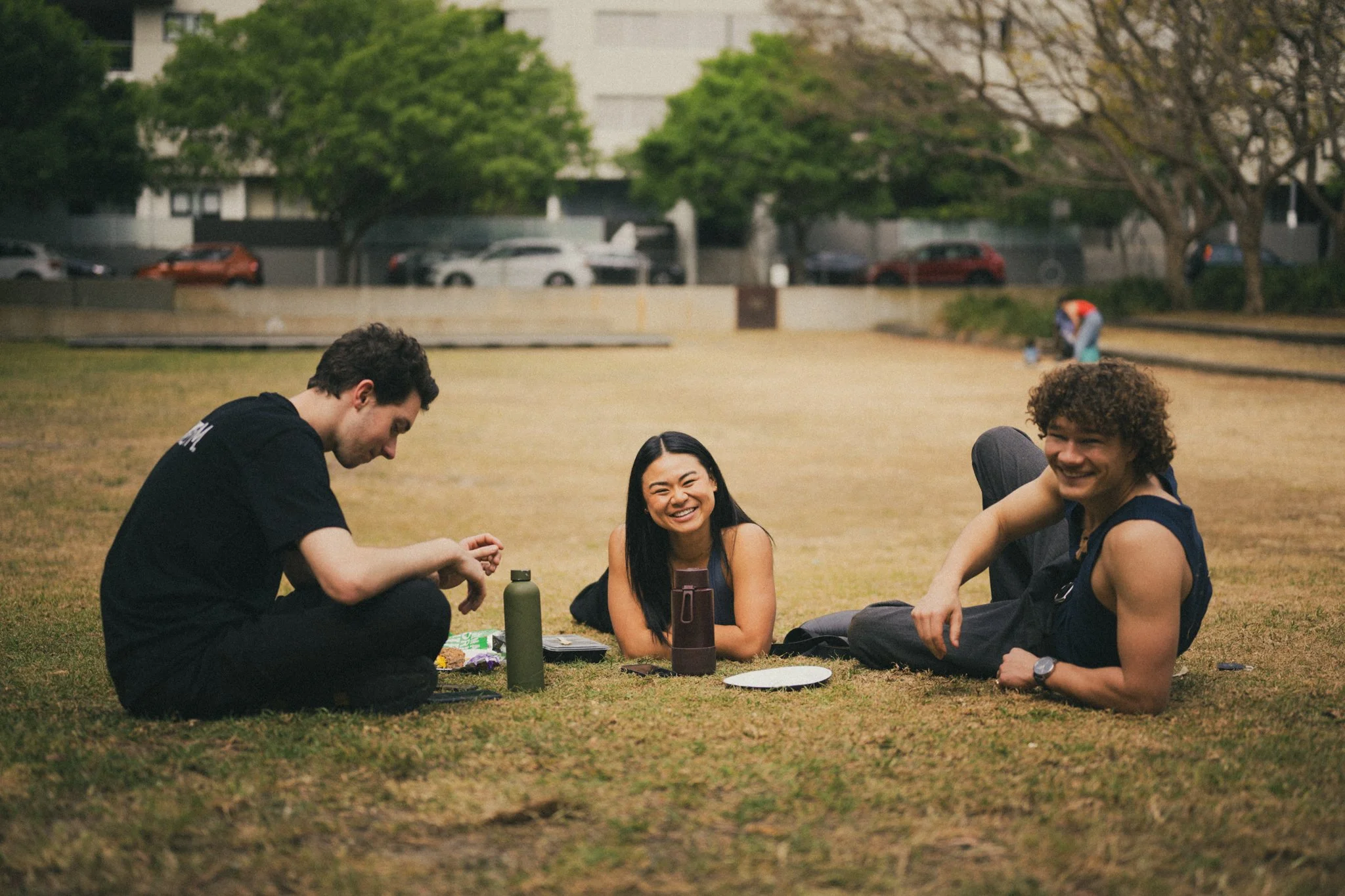 Three friends are relaxing on the grass in a park, smiling and chatting. There are water bottles, snacks, and a plate on the ground.