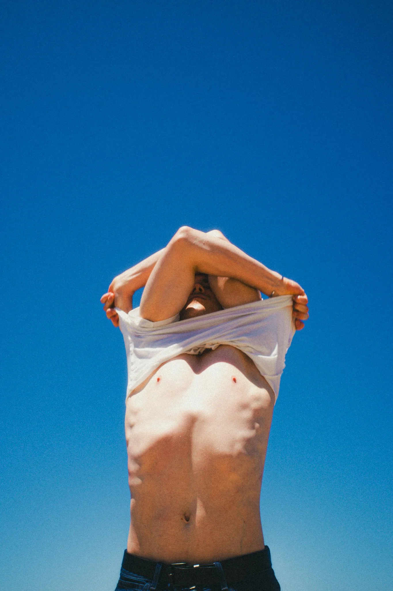 A person with a bare torso is lifting a white shirt over their head against a clear blue sky.