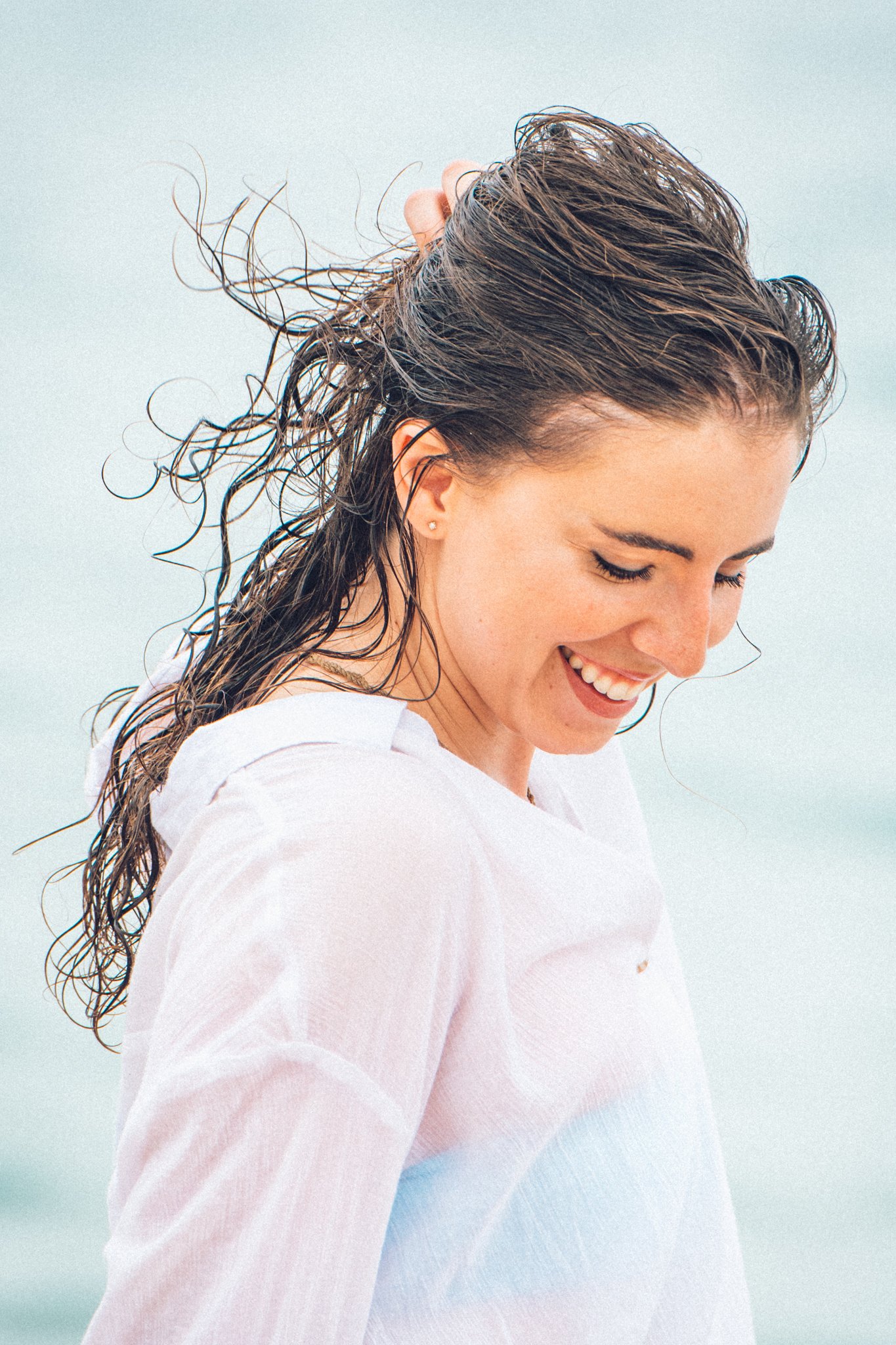 A woman with wet, windblown hair smiling and looking down, wearing a light-colored shirt, outdoors near water.