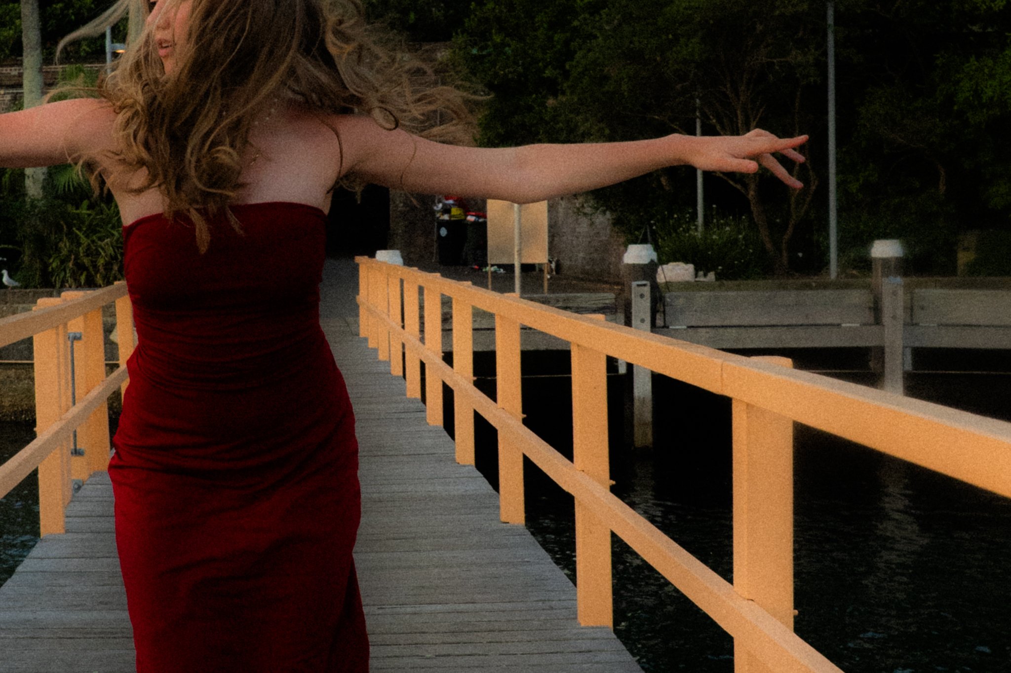 A woman with curly hair wearing a red strapless dress is walking on a wooden dock with her arms outstretched, near water at dusk or evening.