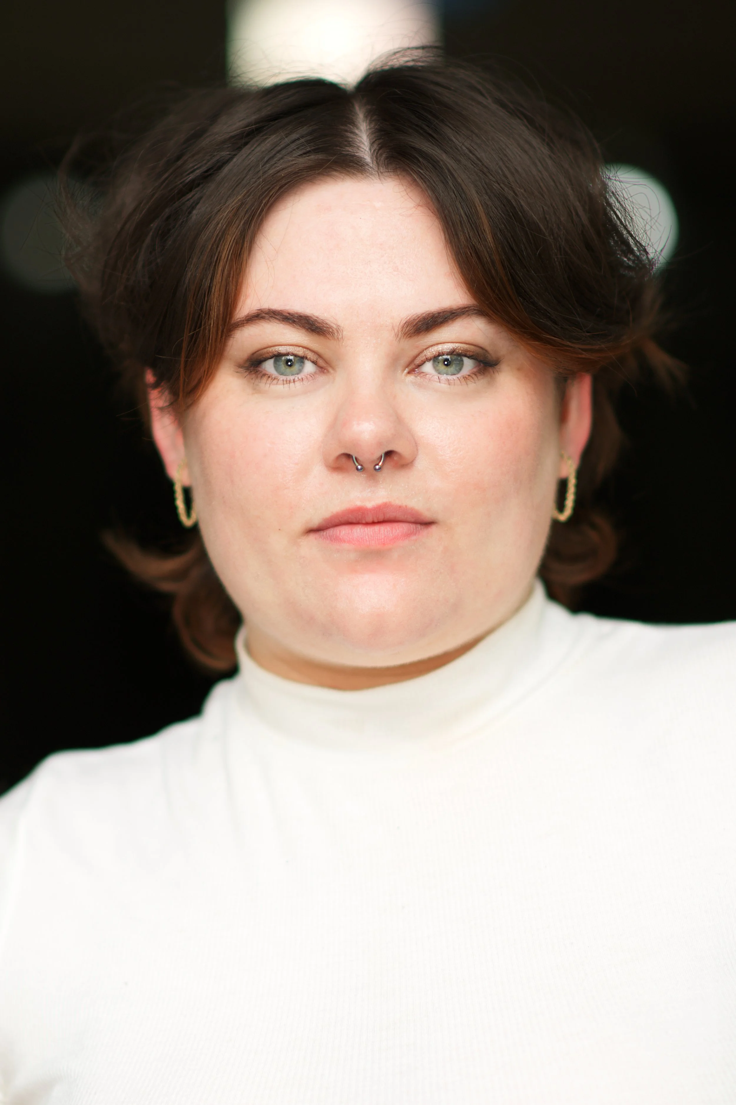 Close-up of a woman with short brown hair and blue eyes, wearing a white turtleneck and gold hoop earrings.