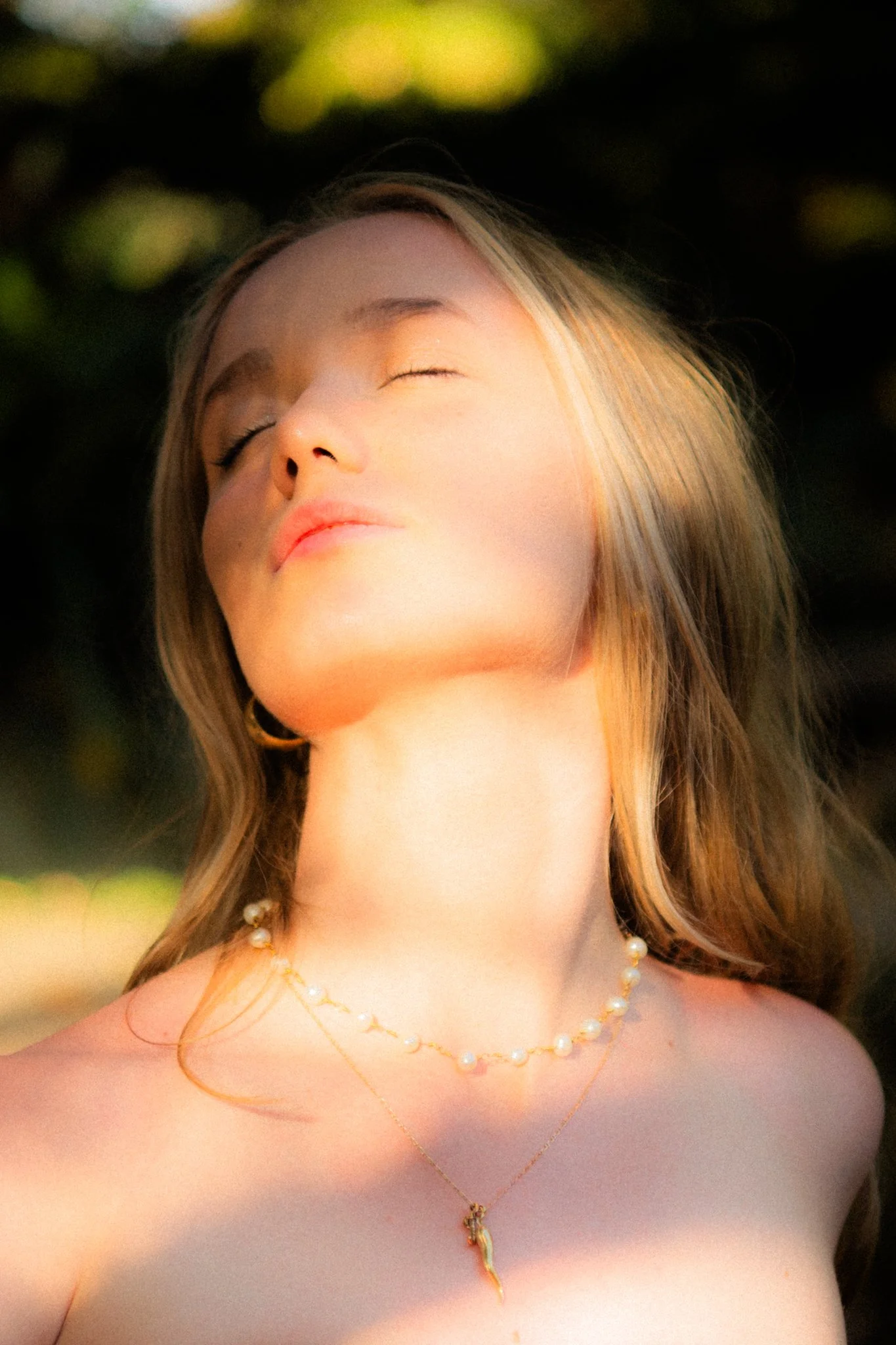 A young woman with light skin and blonde hair, wearing pearl necklaces and gold jewelry, is enjoying the sunlight with her eyes closed and face tilted upward.