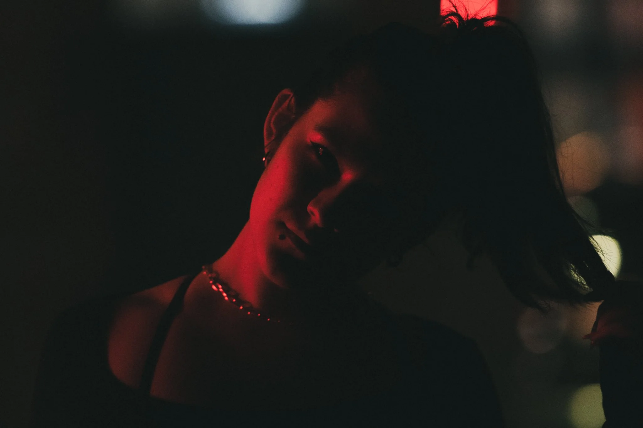 A woman with short hair and earrings, illuminated by red and pink lighting, in a dark setting.