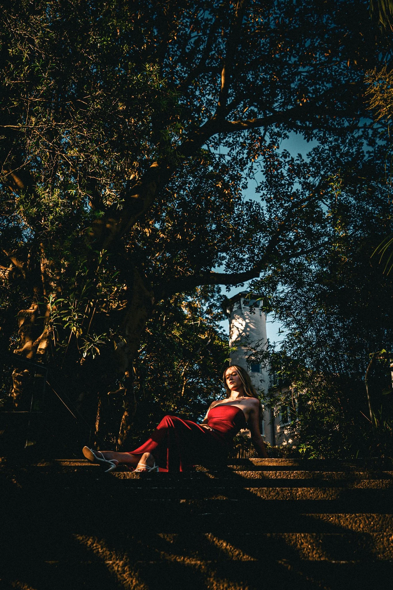 A woman in a red strapless dress sitting on outdoor steps, surrounded by trees, with a lighthouse in the background during sunset.