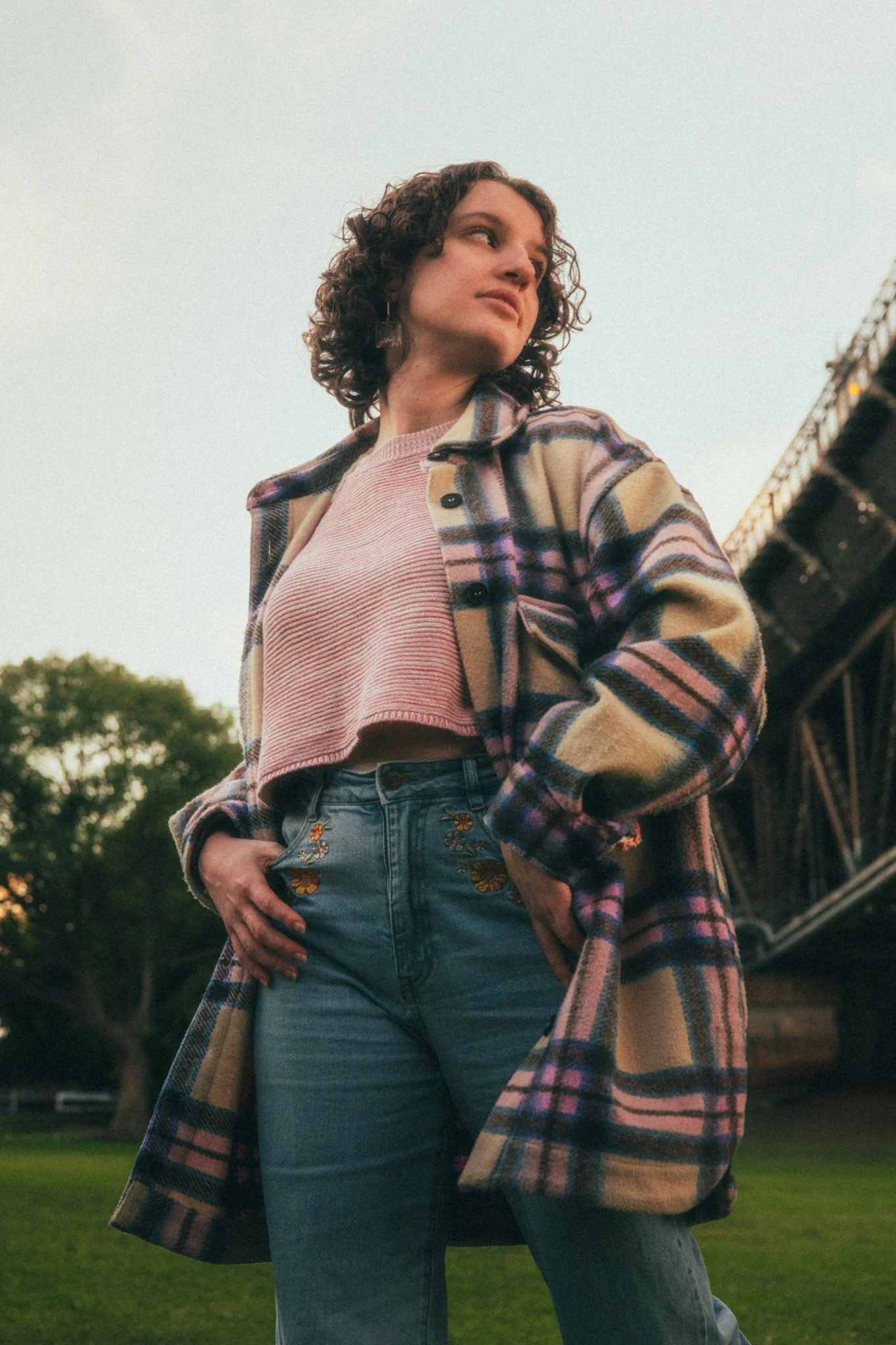 A young woman with curly hair wearing a plaid coat, pink crop top, and embroidered jeans stands outdoors near a bridge and trees during sunset.