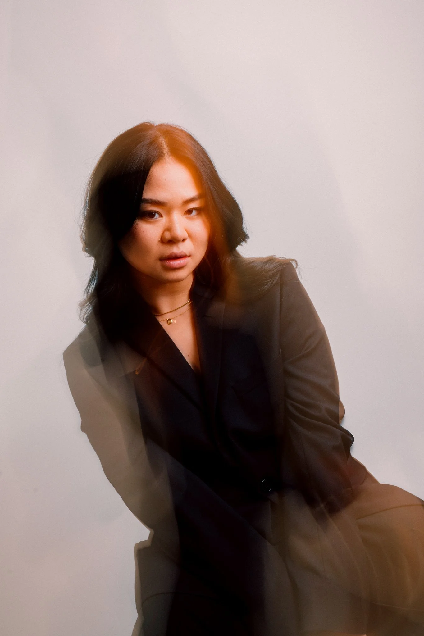 A young woman with dark hair wearing a black blazer, sitting against a plain white background, with a subtle orange light reflection on her face.