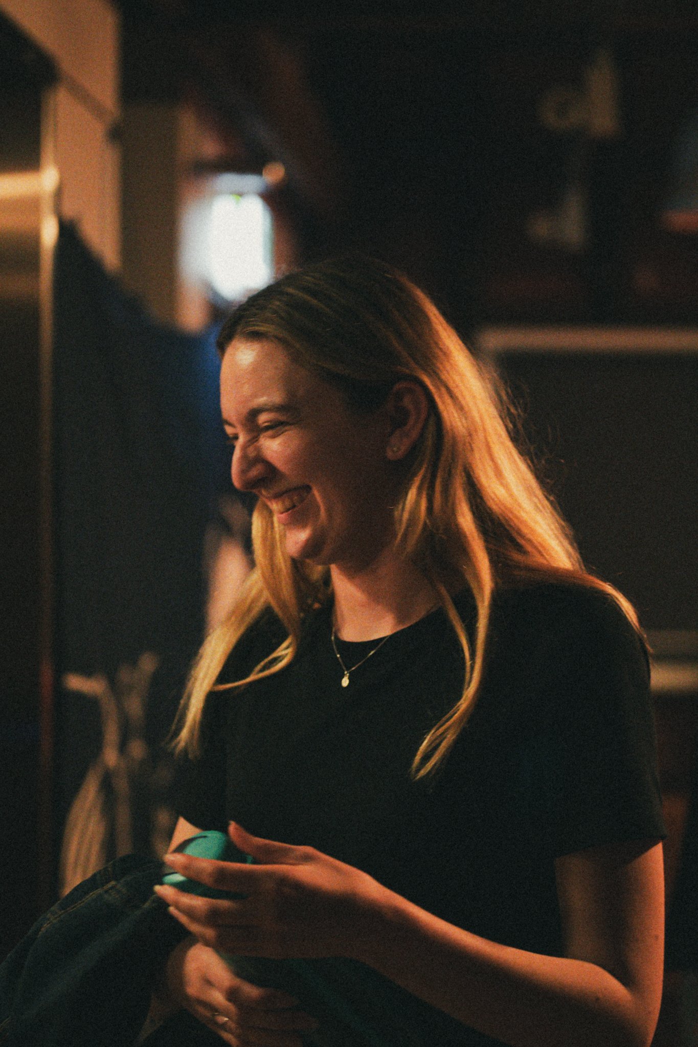 A woman with long blonde hair, smiling and looking down, in a dimly lit indoor setting.