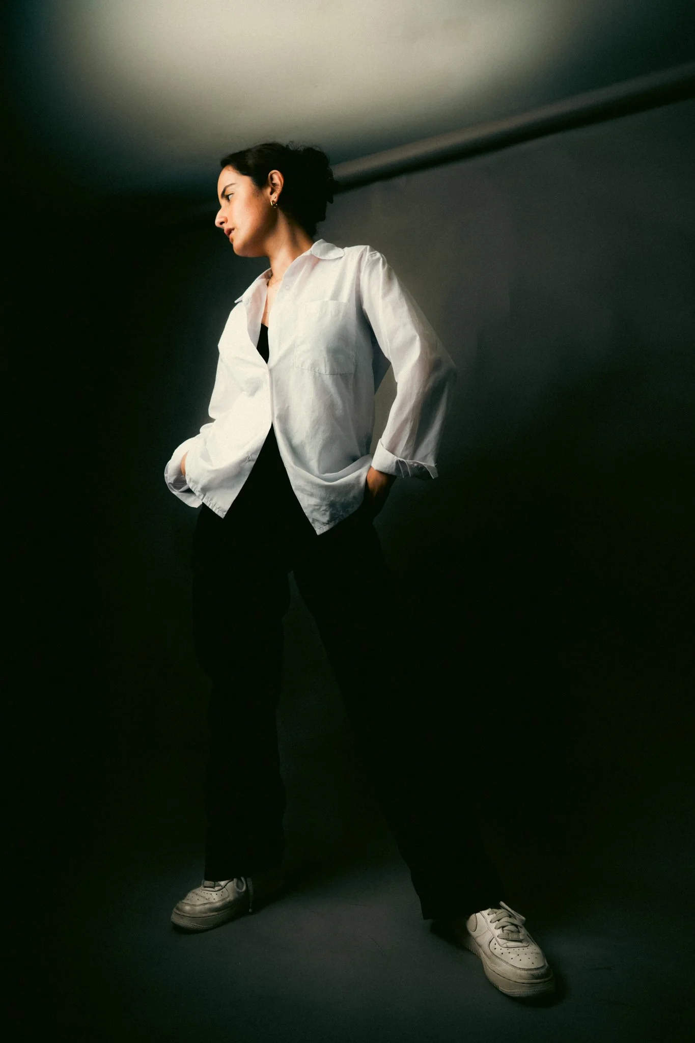 A woman standing against a dark background, wearing a white button-up shirt, black pants, and white sneakers, with hands in her pockets and looking to the side.
