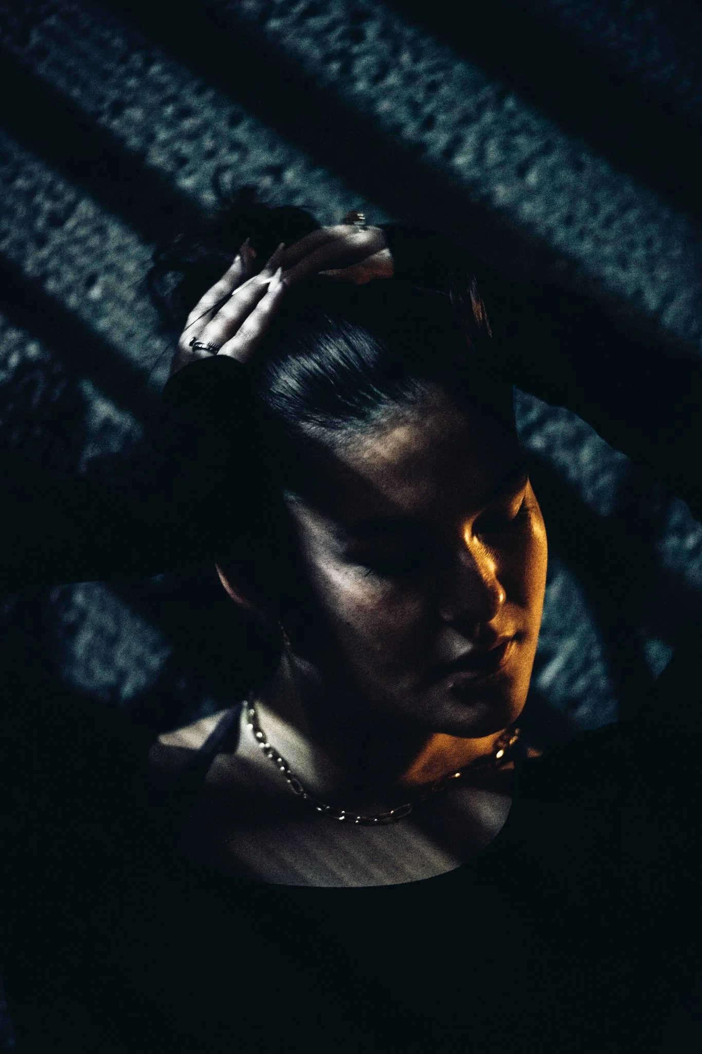 A woman with dark hair, closed eyes, and a silver necklace, holding her head with both hands, illuminated by a single warm light source, creating shadows on her face, against a dark background.
