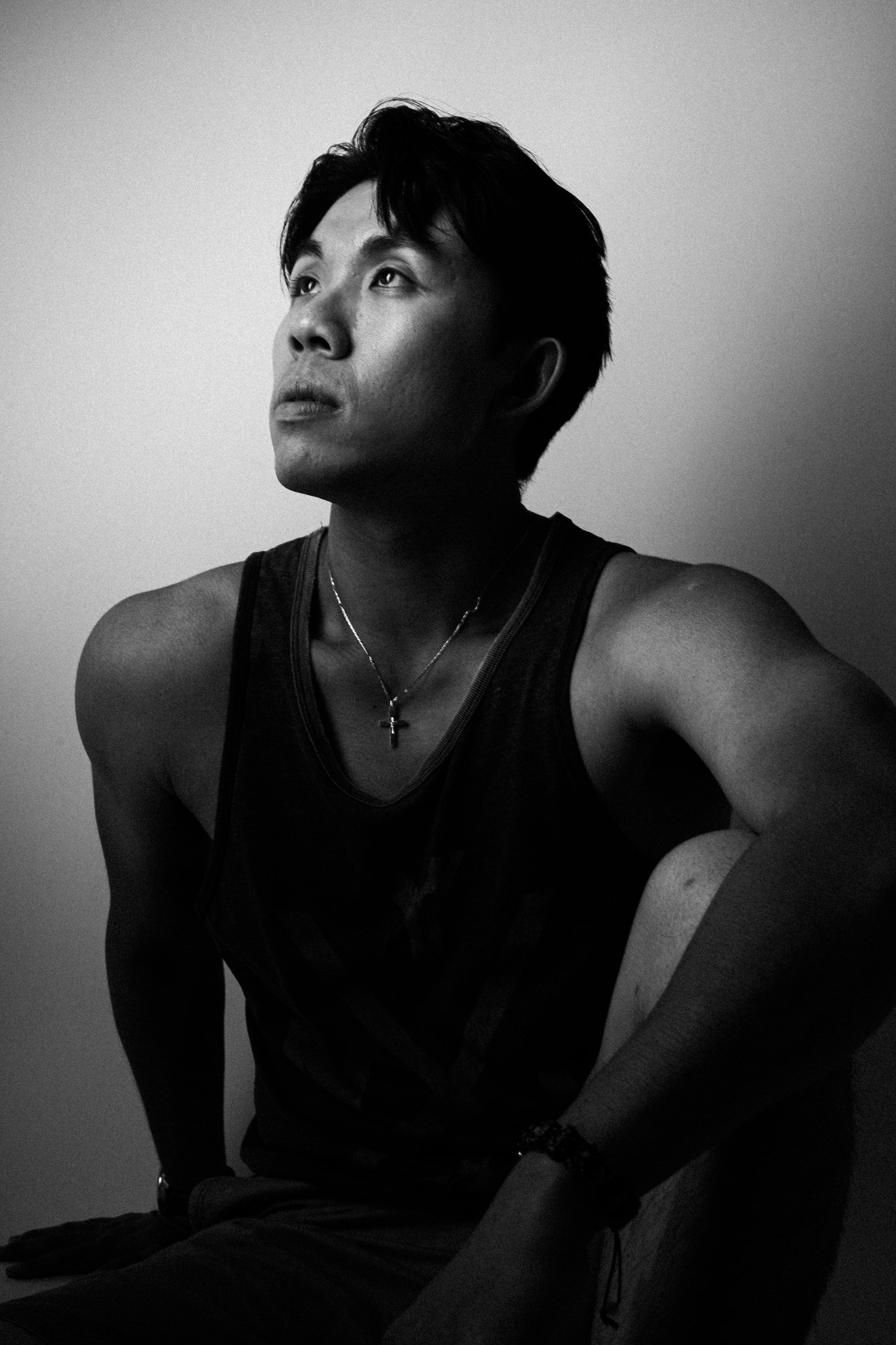 Black and white portrait of a young man with short dark hair, looking upward with a contemplative expression, wearing a tank top, a necklace with a cross, and a bracelet, sitting against a plain background.