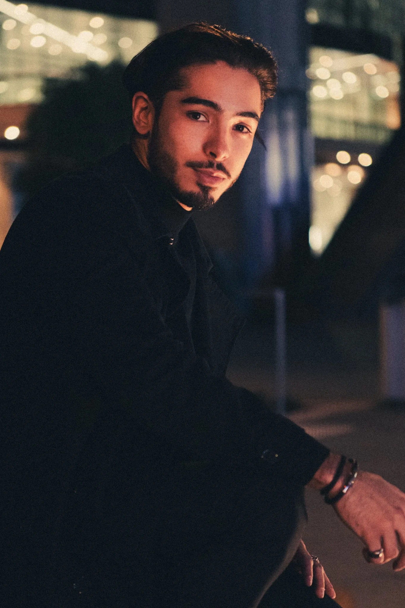 A young man with dark hair and a beard smiling and sitting in an outdoor urban setting at night with blurred lights in the background.