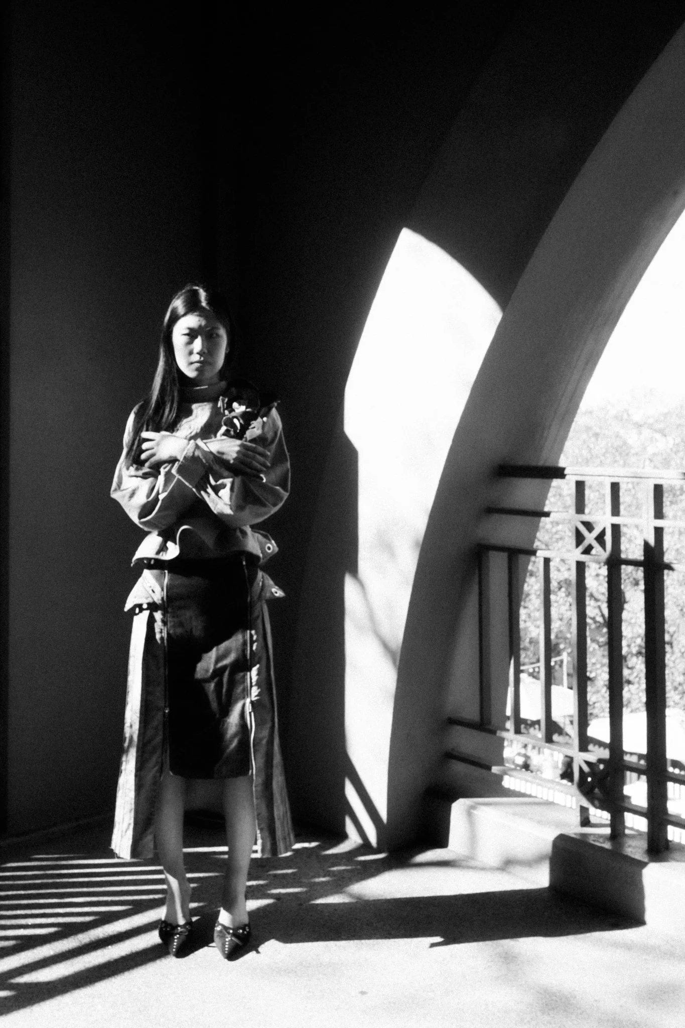 A black-and-white photo of a woman standing near a balcony with sunlight casting shadows. She is looking at the camera, holding a cat, and standing against an arched wall.