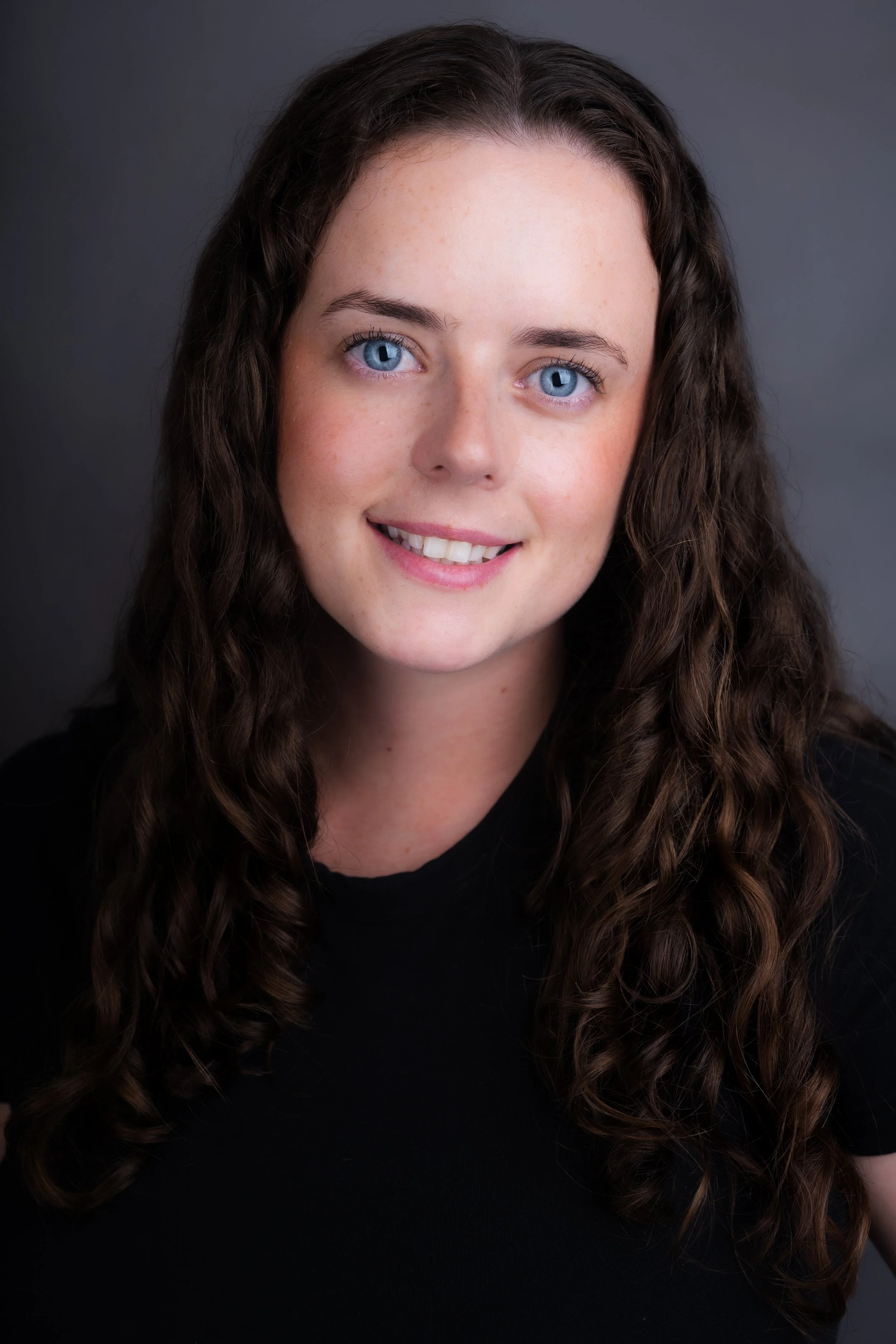 A young woman with long, curly brown hair and blue eyes smiling against a dark gray background.