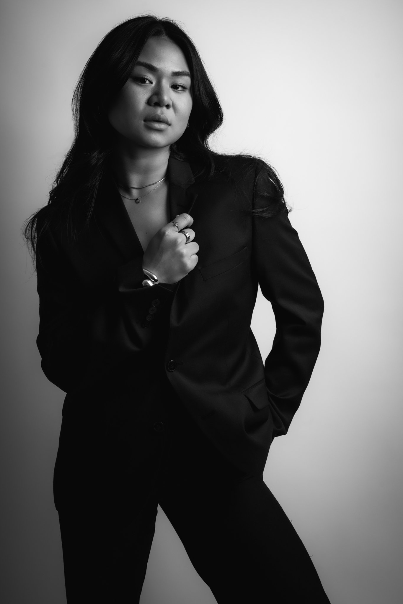 Black and white photo of a woman with long hair, wearing a dark suit, with her right hand holding the lapel of her jacket. She has a confident expression, with several rings and layered necklaces.