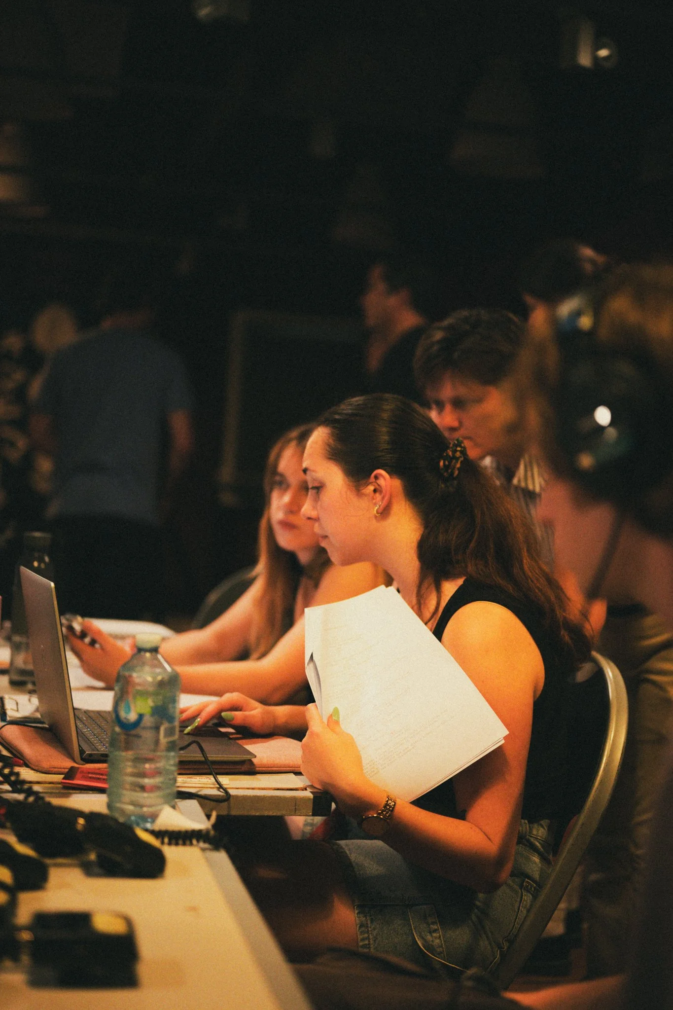 A group of young women sitting at a table working on laptops, reviewing documents, in a dimly lit environment, possibly a conference or workshop.