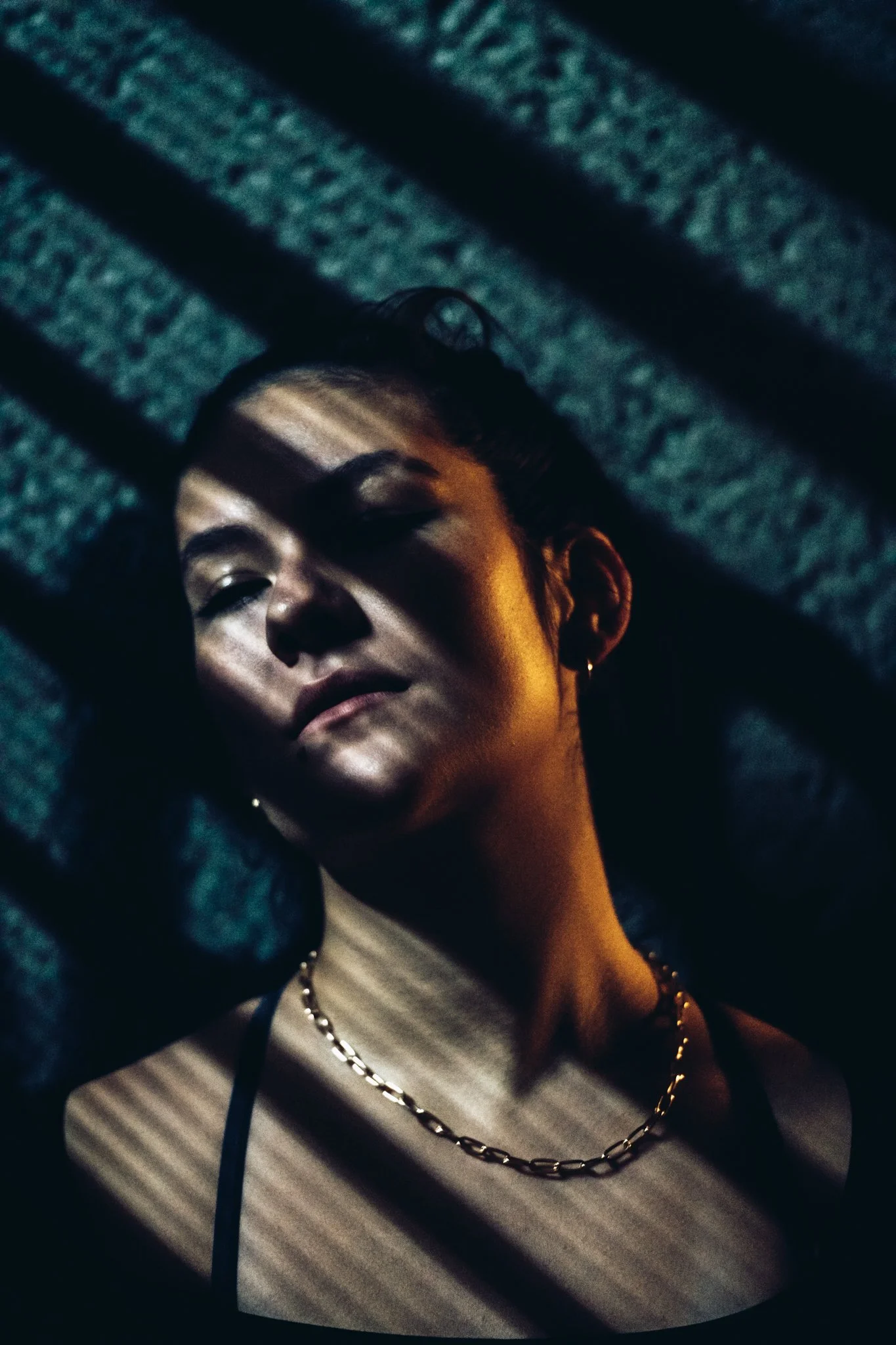A woman with dark hair and earrings lies on a textured surface, partially obscured by shadows cast by striped light patterns.