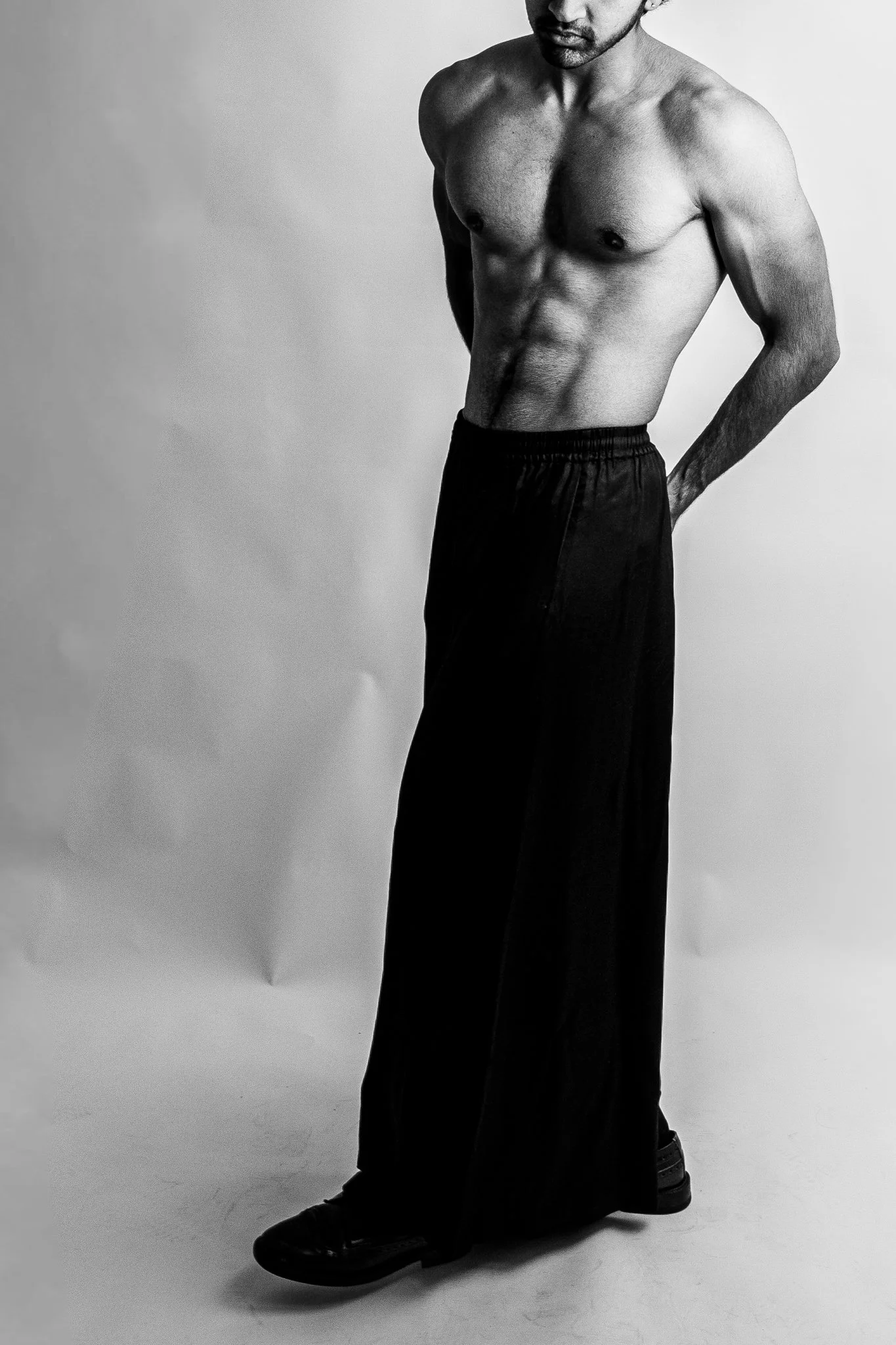 Black and white photo of a shirtless man with muscular build wearing dark loose pants and shoes, standing against a plain background.