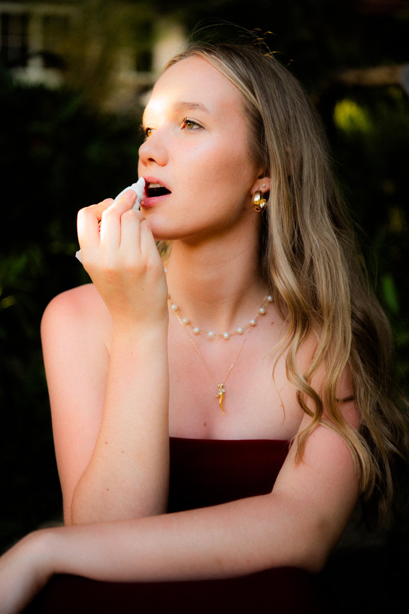 A young woman with long wavy hair and gold jewelry, holding a lip balm near her lips outdoors.