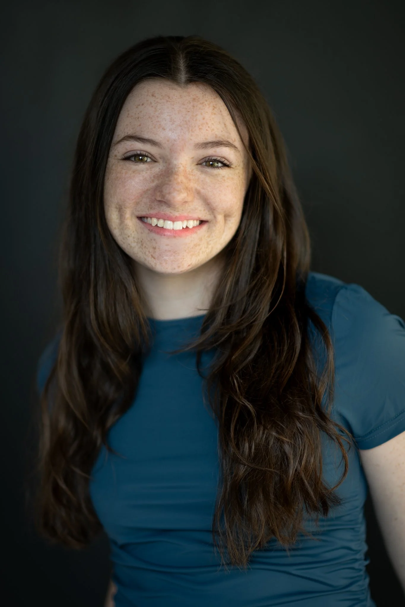 Portrait of a young woman with long dark hair, light skin with freckles, smiling, wearing a blue shirt, against a dark background.